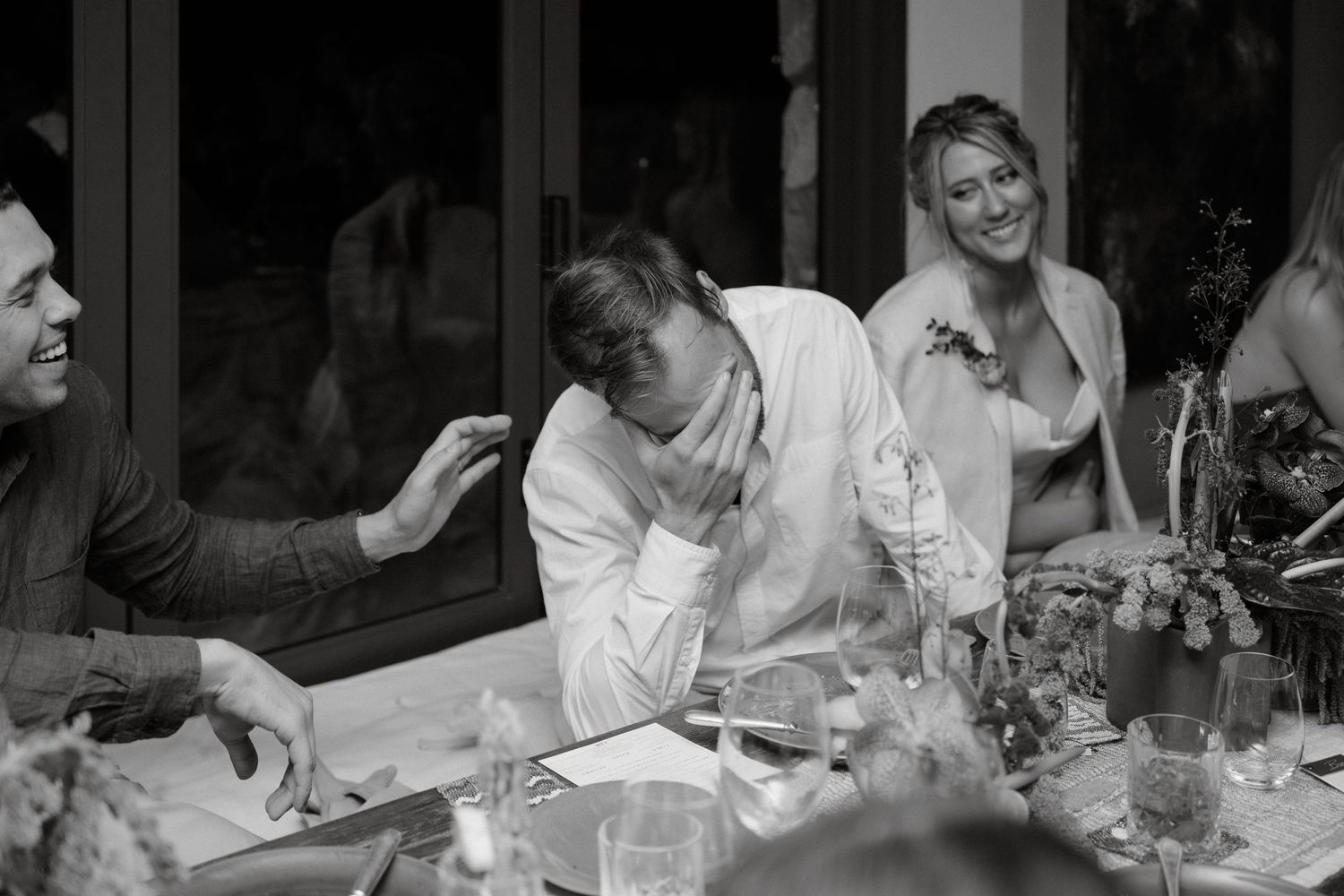 A black and white photo of guests sharing laughter and conversation around a dinner table at an event.