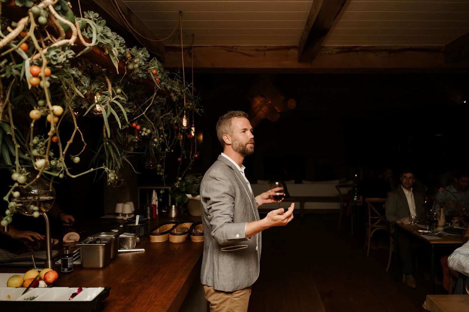 Guest in grey blazer giving toast at rustic venue with hanging greenery.