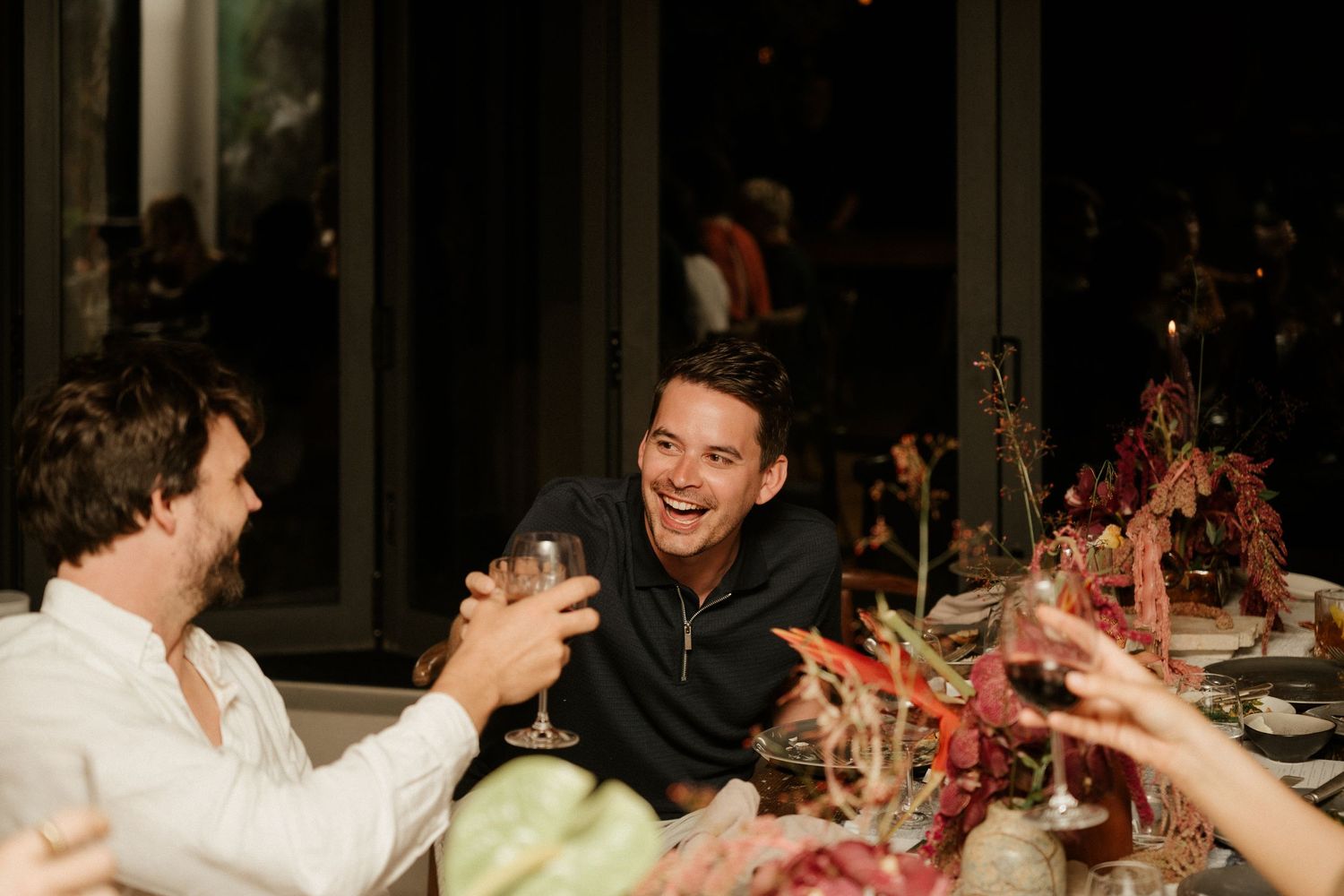 Guests laugh and celebrate at dimly lit wedding reception dinner table.