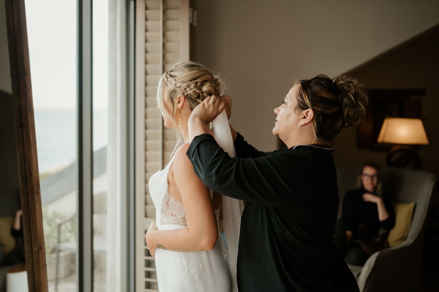 A touching moment during wedding preparations as final adjustments are made near a bright window.