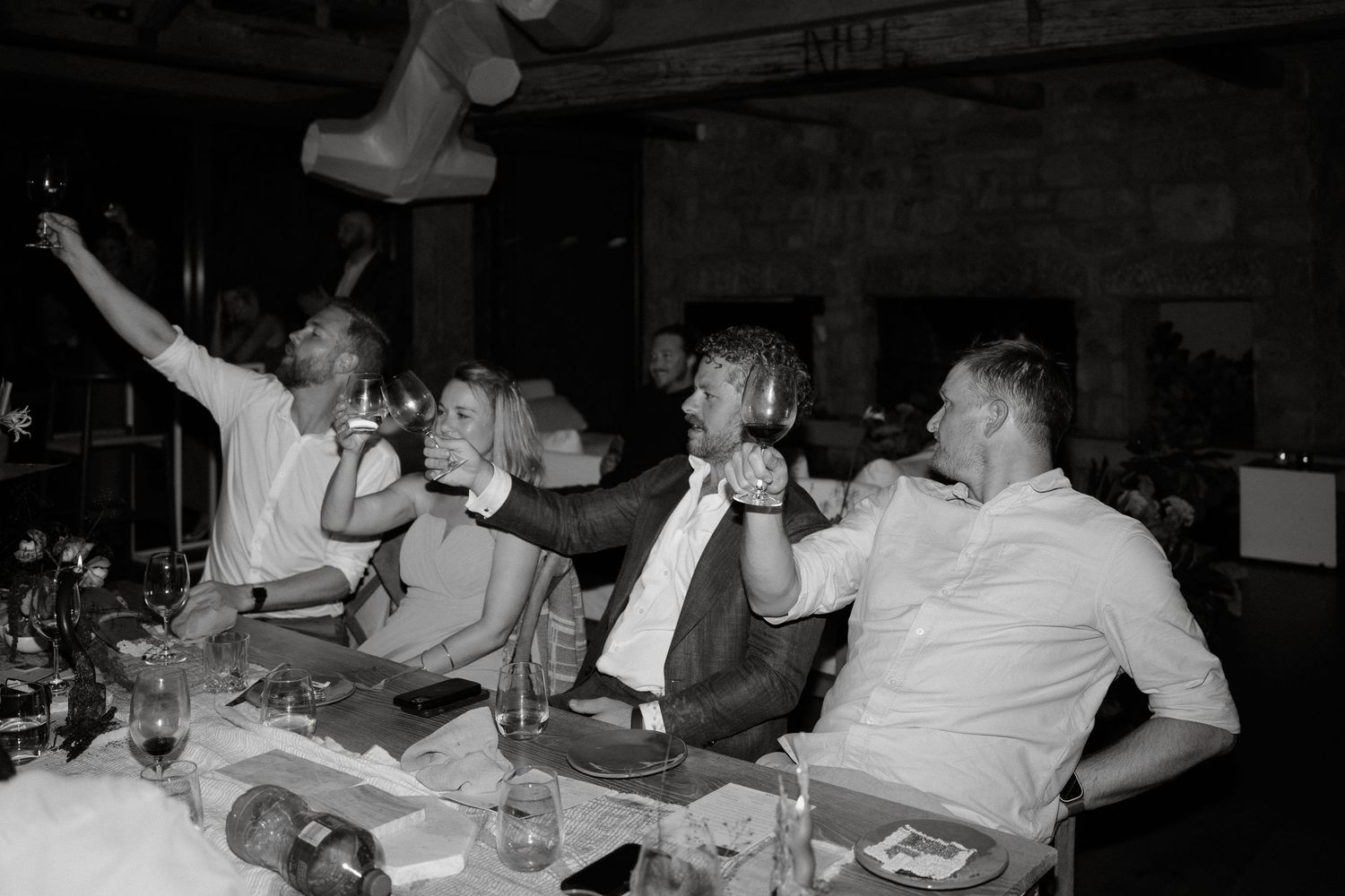 Black and white candid shot of wedding guests enjoying drinks and raising their glasses in celebration.