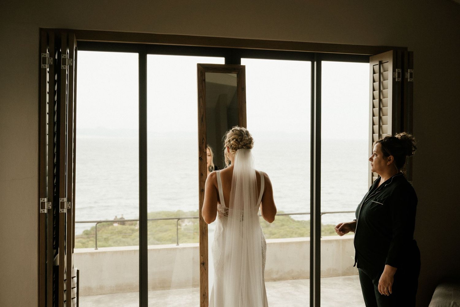 Two people stand in an elegant room with floor-to-ceiling windows overlooking an oceanfront view.