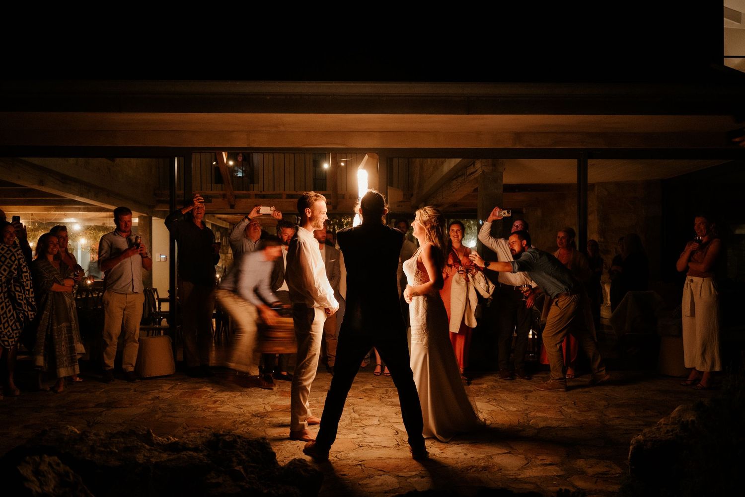 Silhouetted couple dancing in rustic barn venue with ambient lighting and guests in background.