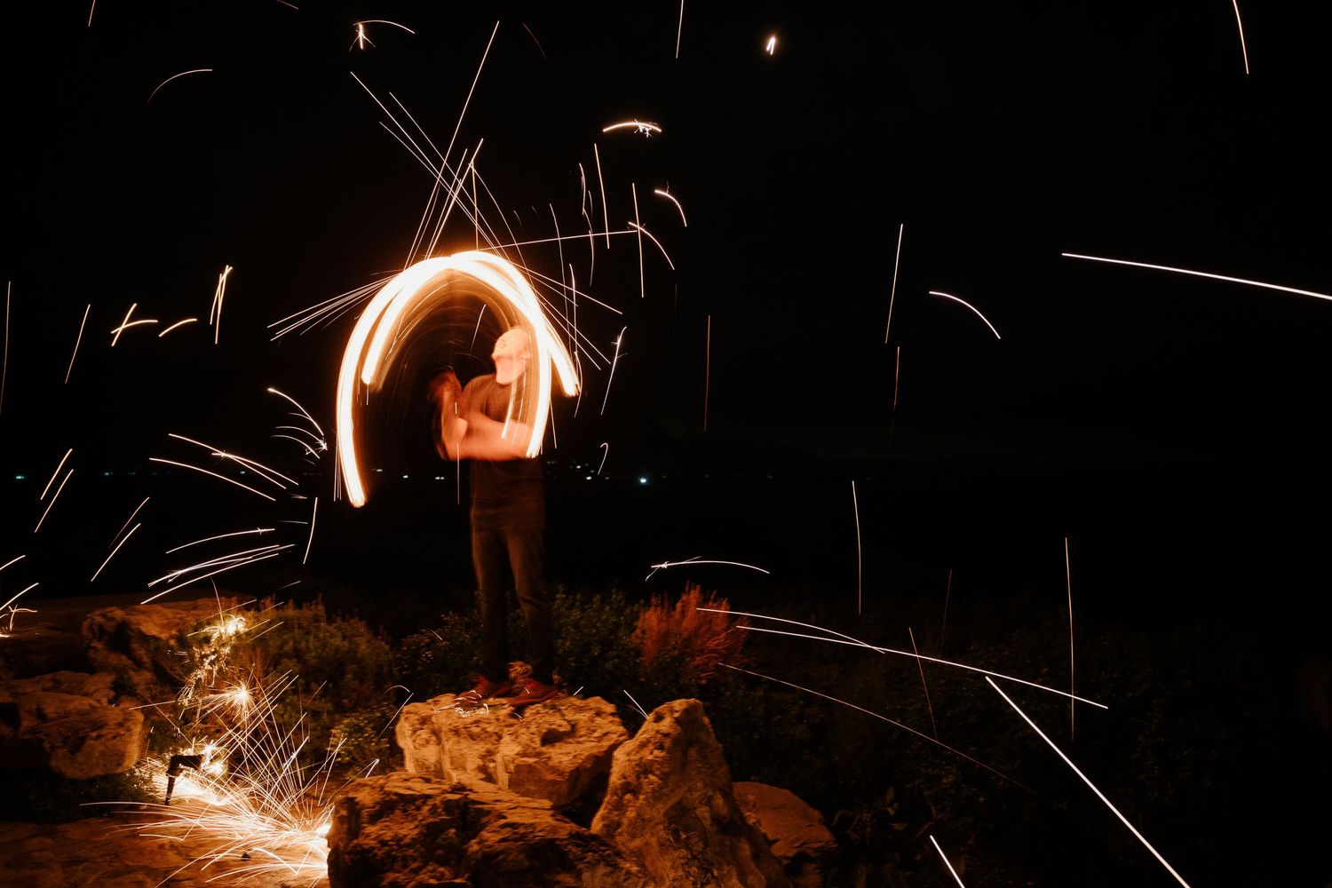 Person creates light painting with sparklers against dark night sky on rocky terrain.