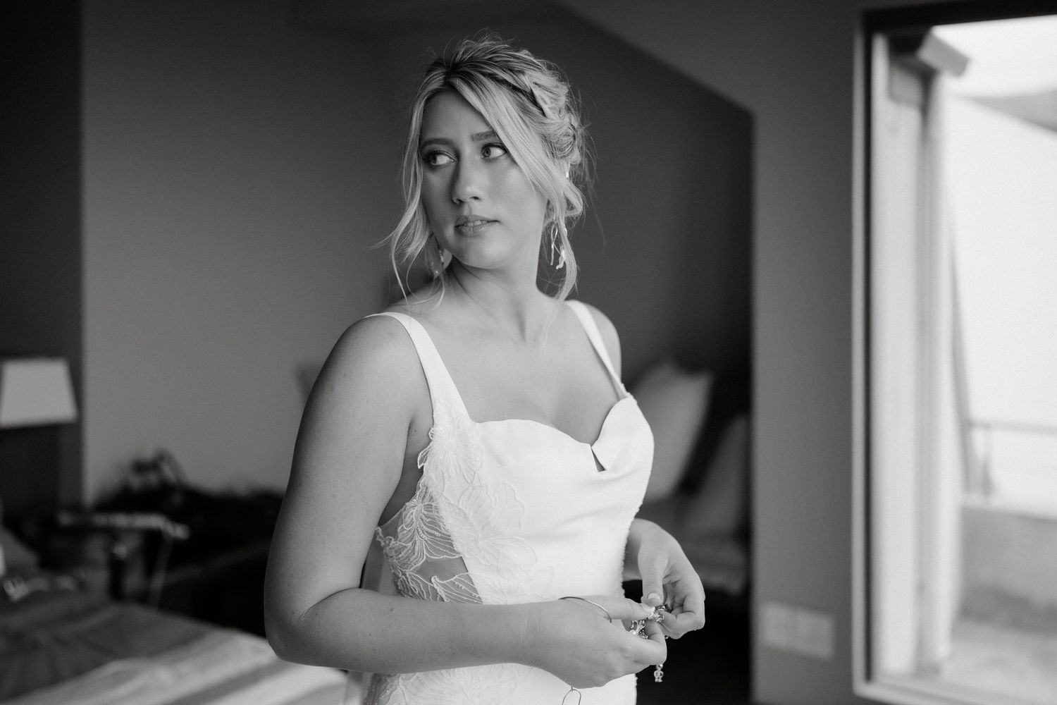 Black and white portrait of a woman in a white wedding dress looking pensive near a window.
