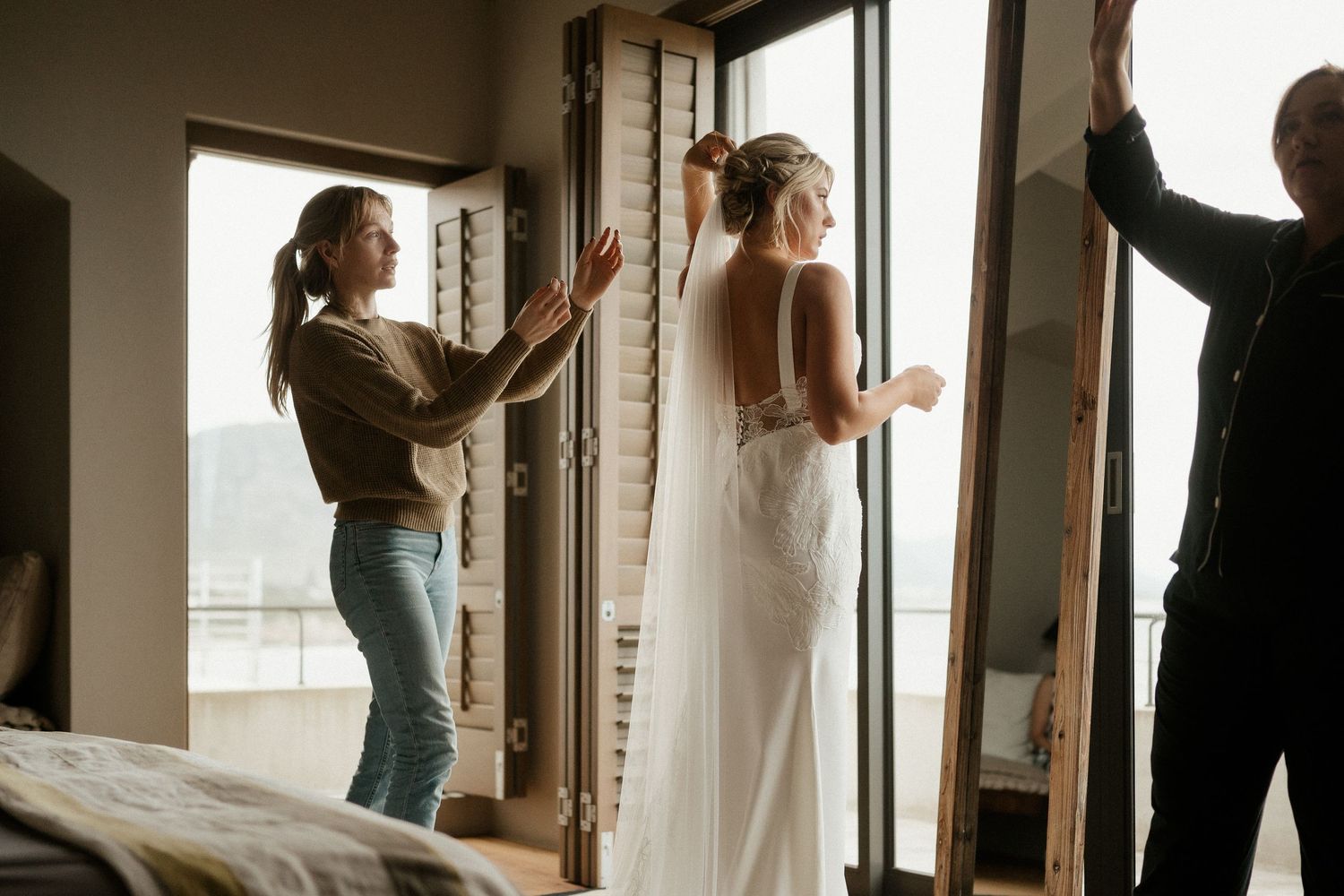 A bride in a flowing white dress gets ready with assistance by a window in a sunlit room.