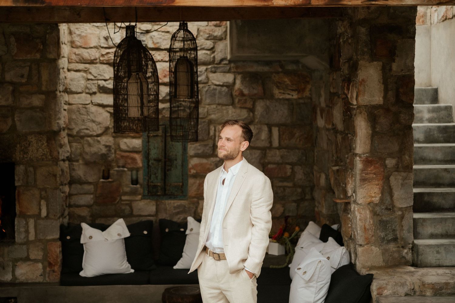 A person in a beige suit waits in an elegant stone archway with antique lanterns and pillows.