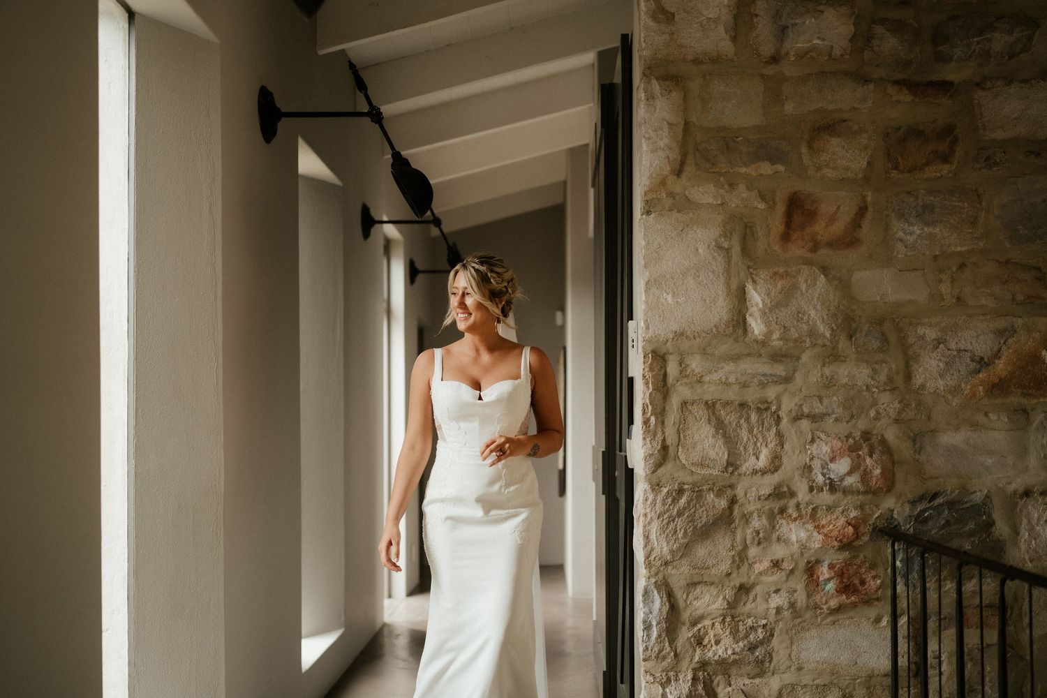 A person in a white wedding dress stands near sunlit windows in a stone hallway.