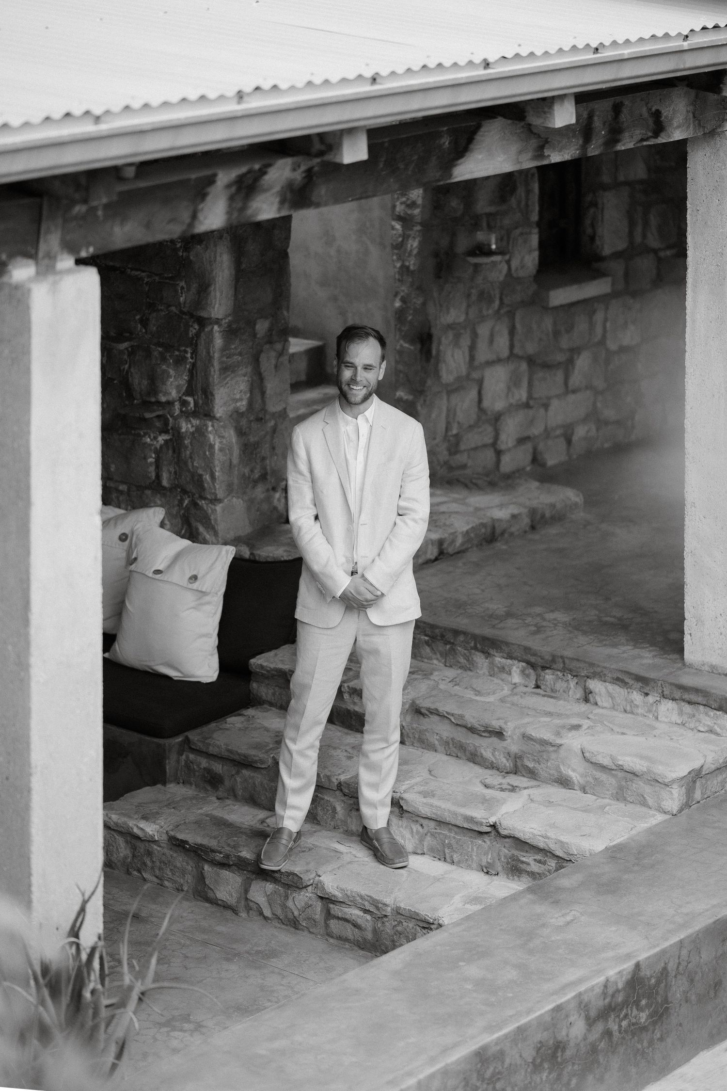 A person in a light colored suit stands on stone steps against a rustic wall.