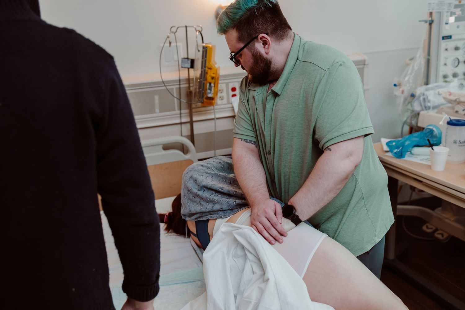 A healthcare worker in green scrubs provides medical care to a patient in a hospital bed.