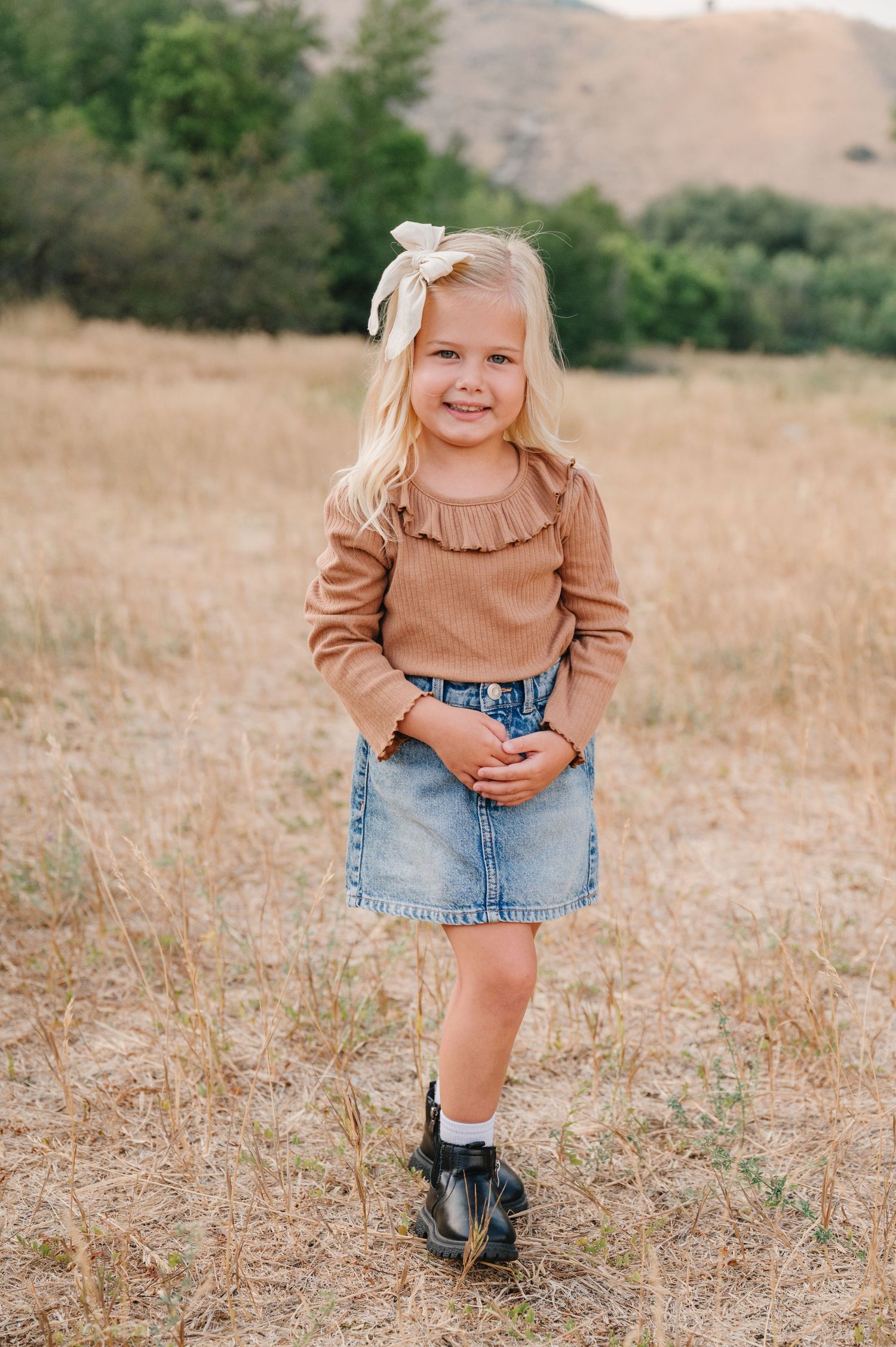 A child wearing a brown sweater and denim shorts stands in a golden field on a sunny day.