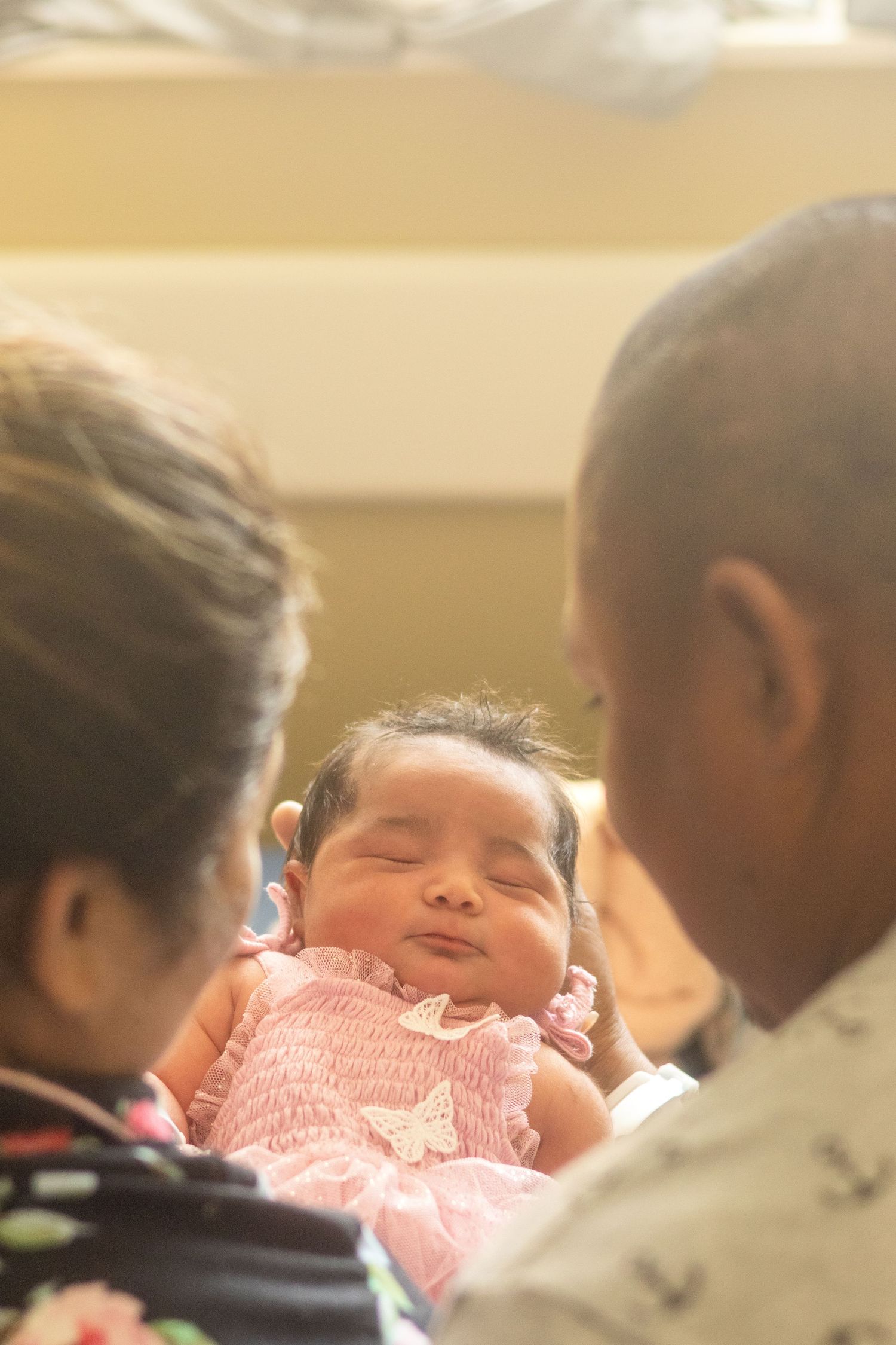 A small baby in a pink outfit smiles while being held by family members.
