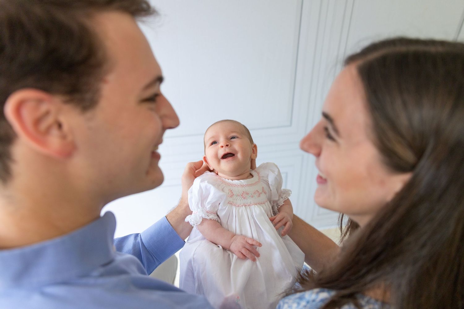 Happy baby in white dress laughing between smiling parents during a bright family portrait session.