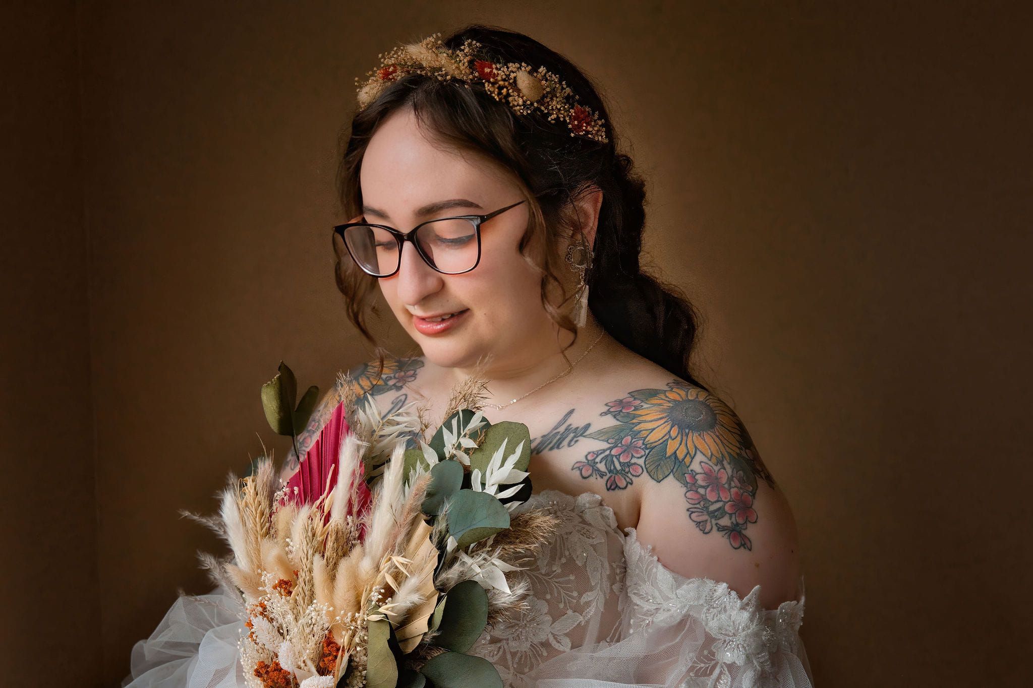 A bride in glasses and lace dress holds a dried flower bouquet while wearing a delicate floral crown.