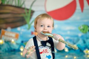 Toddler in blue overalls holding a large spoon against a colorful underwater-themed backdrop.