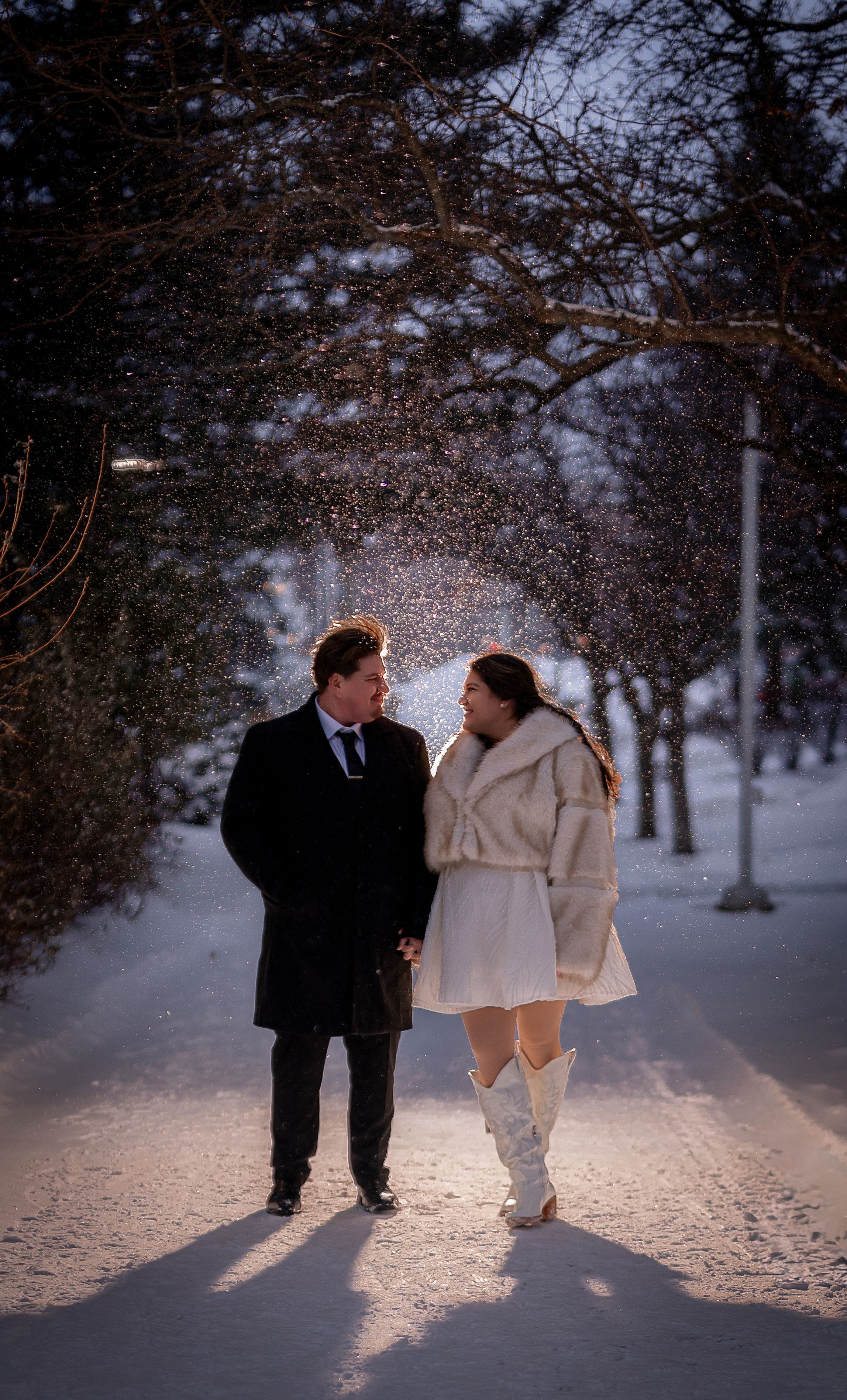 A couple walks together on a snowy path surrounded by winter trees at dusk, wearing elegant winter coats.