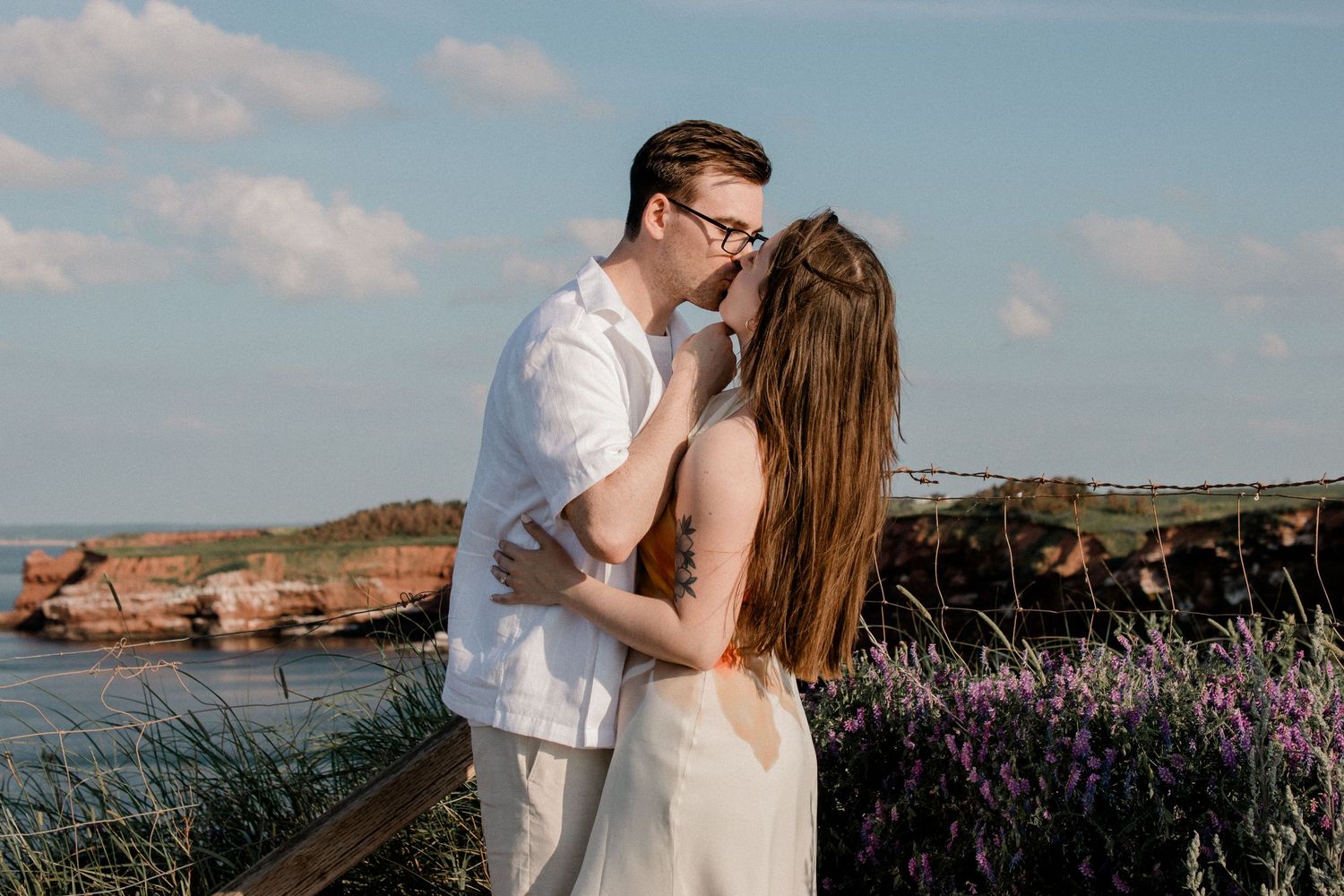 A romantic couple embraces near coastal cliffs with purple wildflowers and ocean views at sunset.