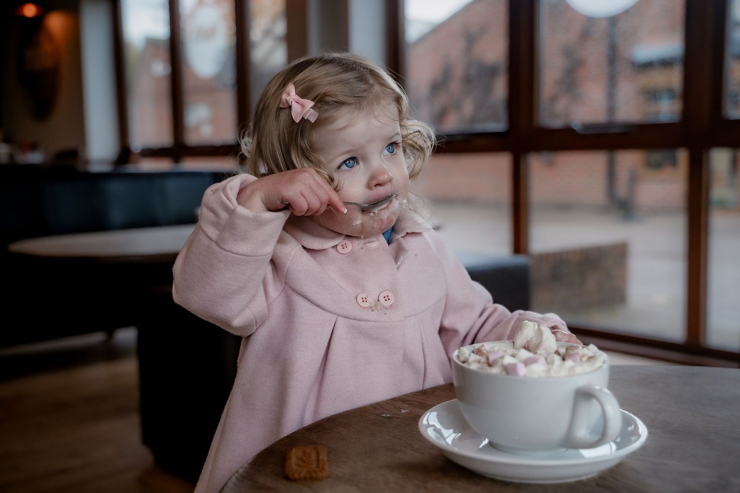 A person in a pink coat enjoys a cozy hot chocolate with marshmallows in a cafe setting.