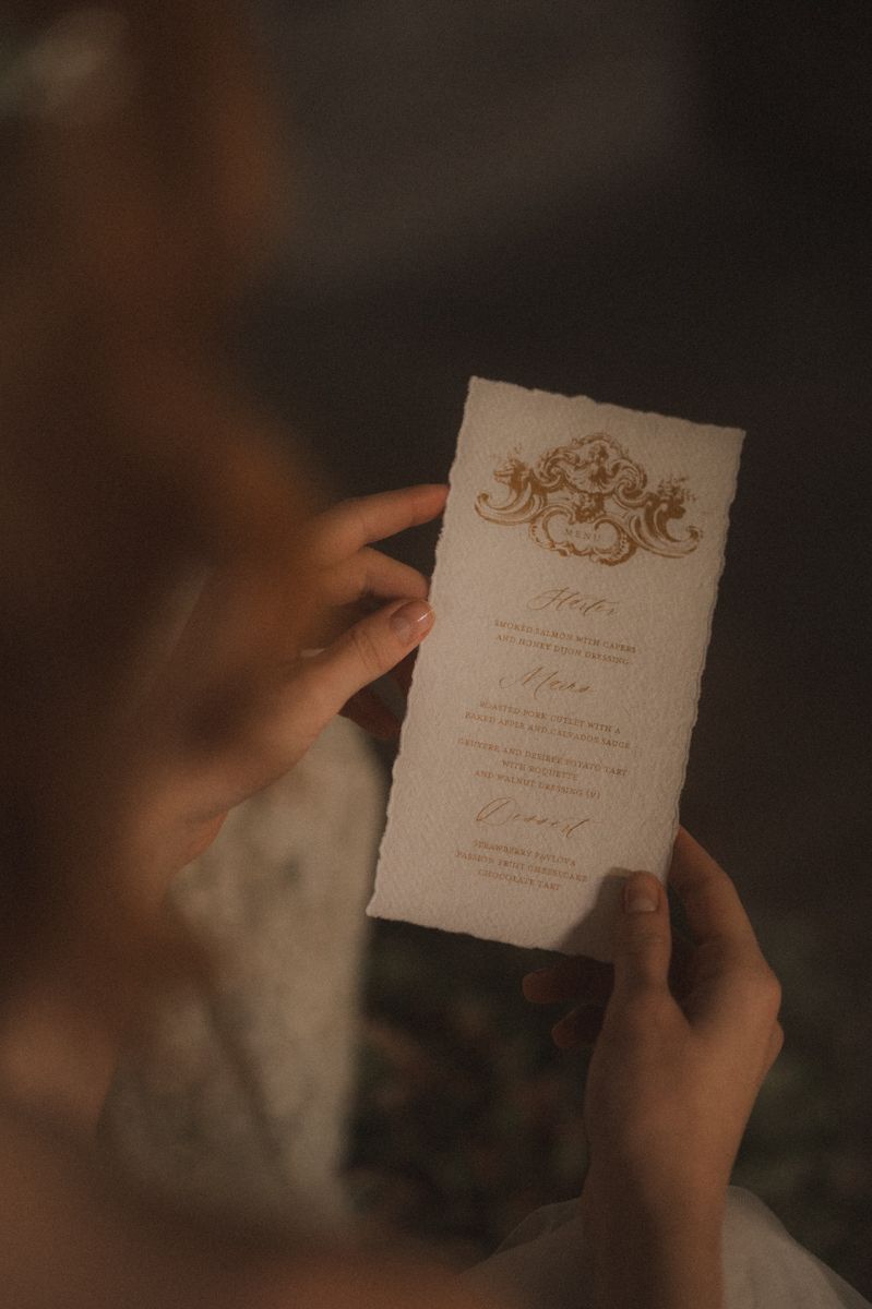 A hand holds an elegant wedding invitation with gold script lettering and ornate details in warm lighting.