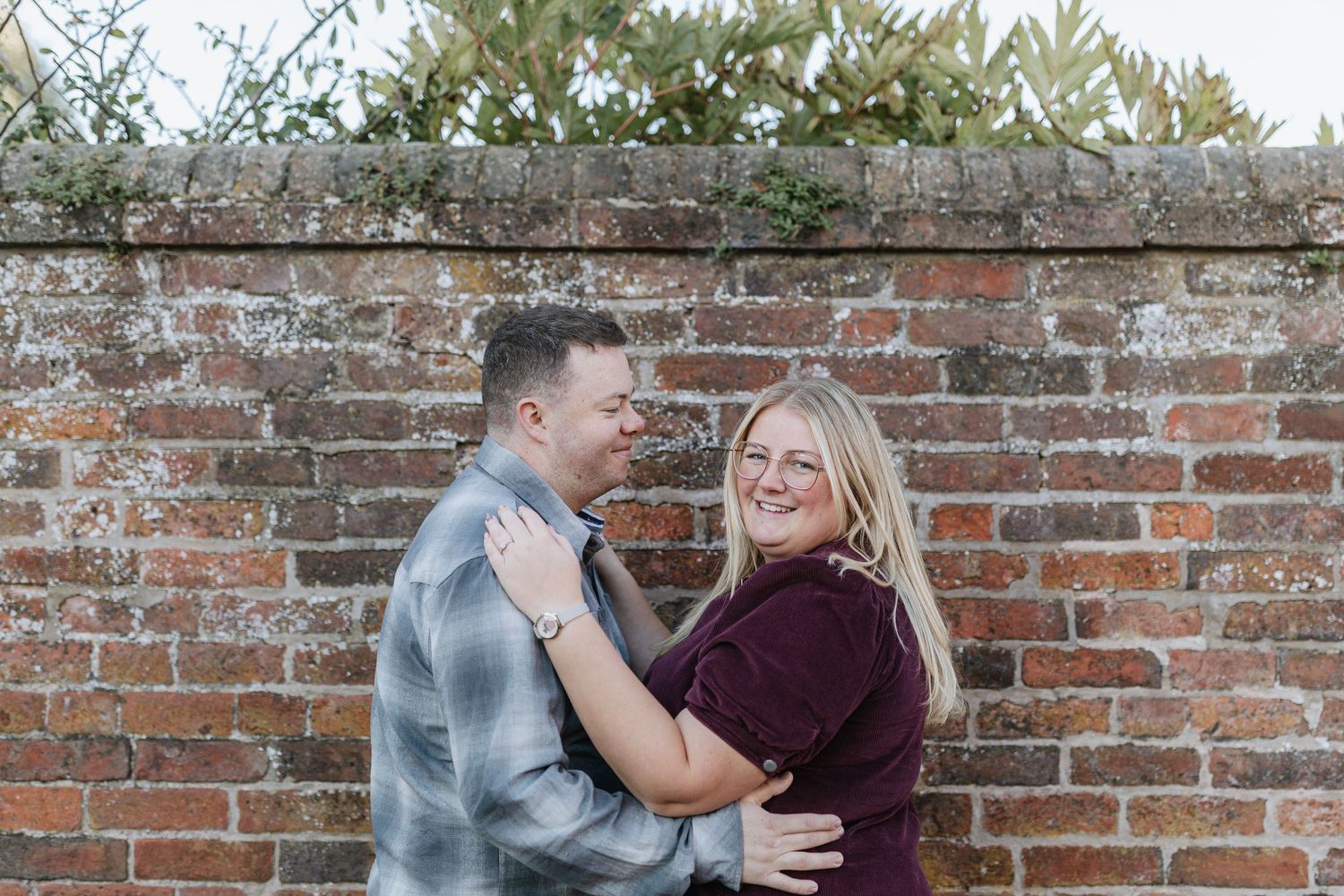 A couple shares a romantic moment against a rustic brick wall backdrop during an outdoor photoshoot.