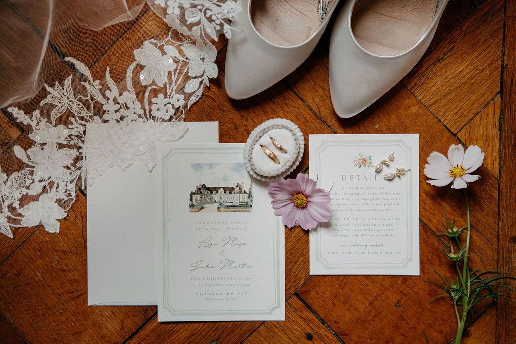 Flat lay of wedding stationery, lace veil, bridal shoes, earrings, and a fresh cosmos flower on wood.