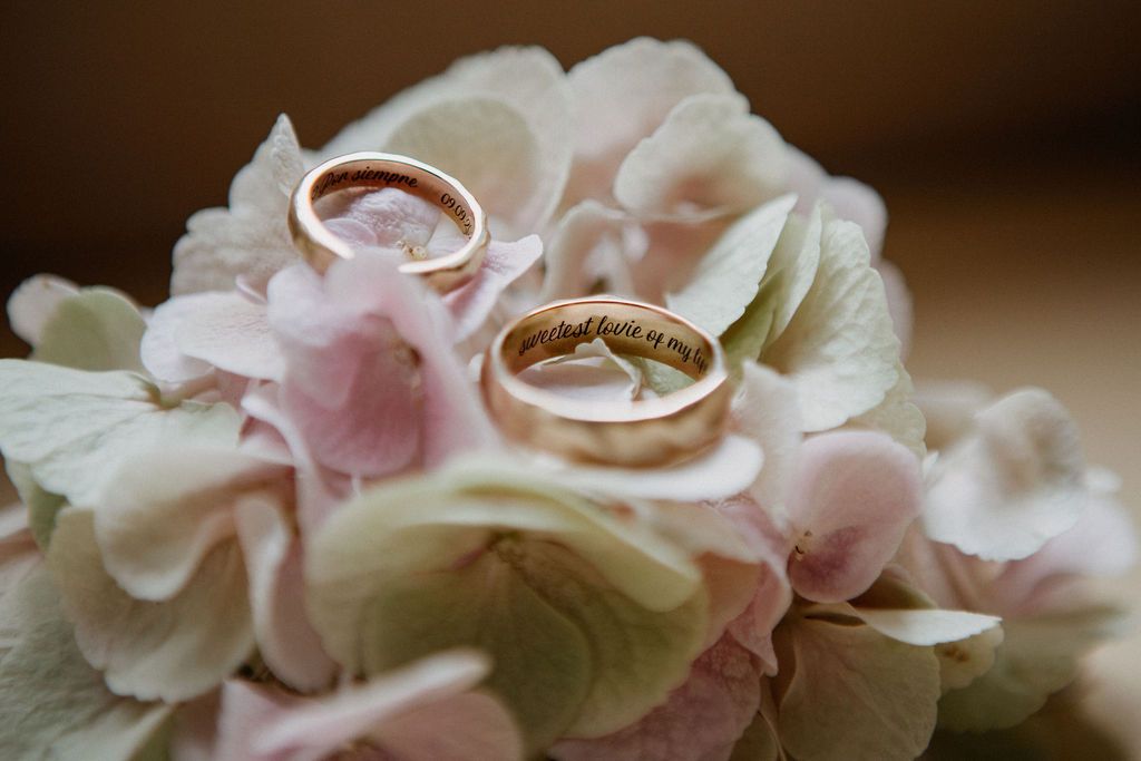 Two gold wedding bands resting on soft pink and white hydrangea flowers with engraved inscriptions.