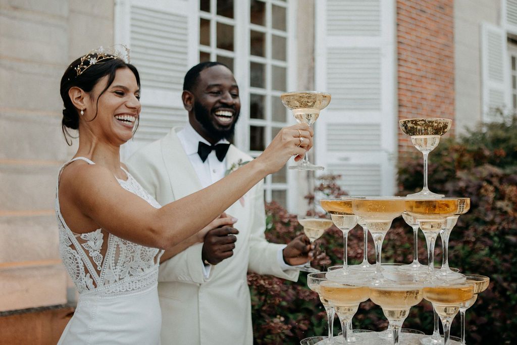 Laughing bride and groom toasting with champagne glasses from an overflowing champagne tower outdoors.