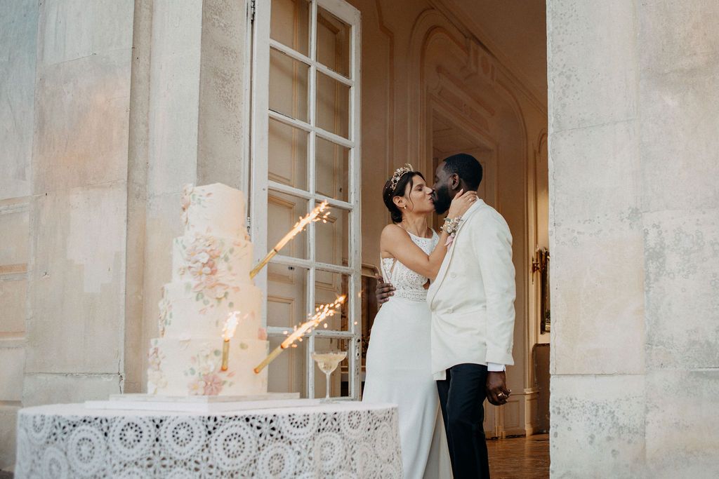 Bride and groom sharing a romantic kiss beside a sparkling wedding cake with sparklers at a chateau.