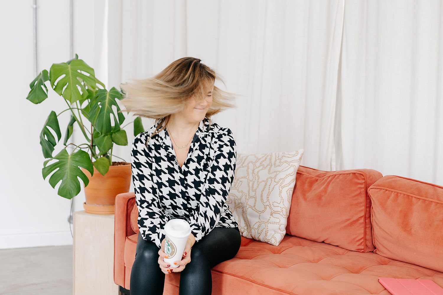 Person in black and white houndstooth top sits on coral couch with monstera plant in modern interior setting.