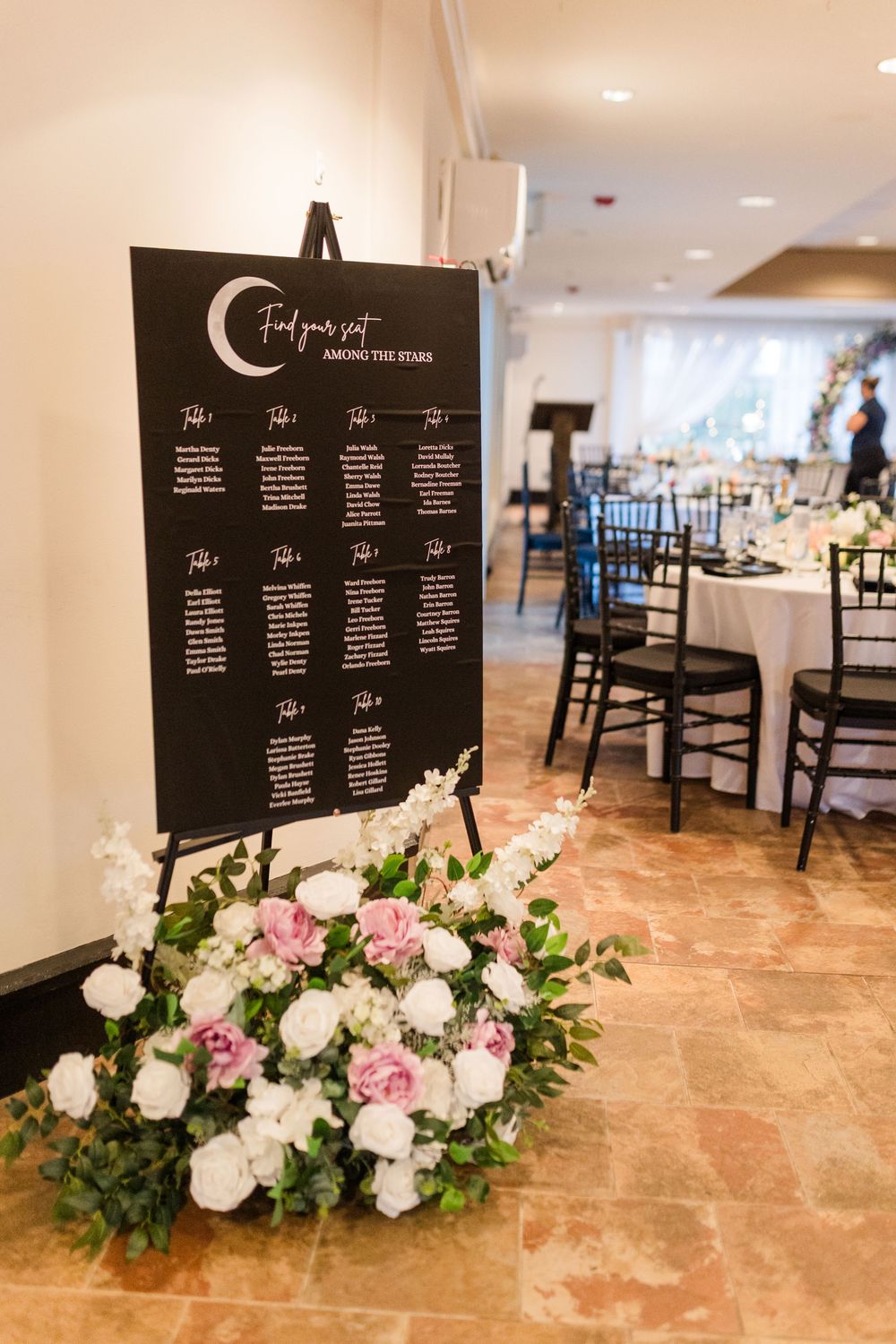Wedding reception seating chart display with floral arrangements featuring pink and white roses on a marble floor.