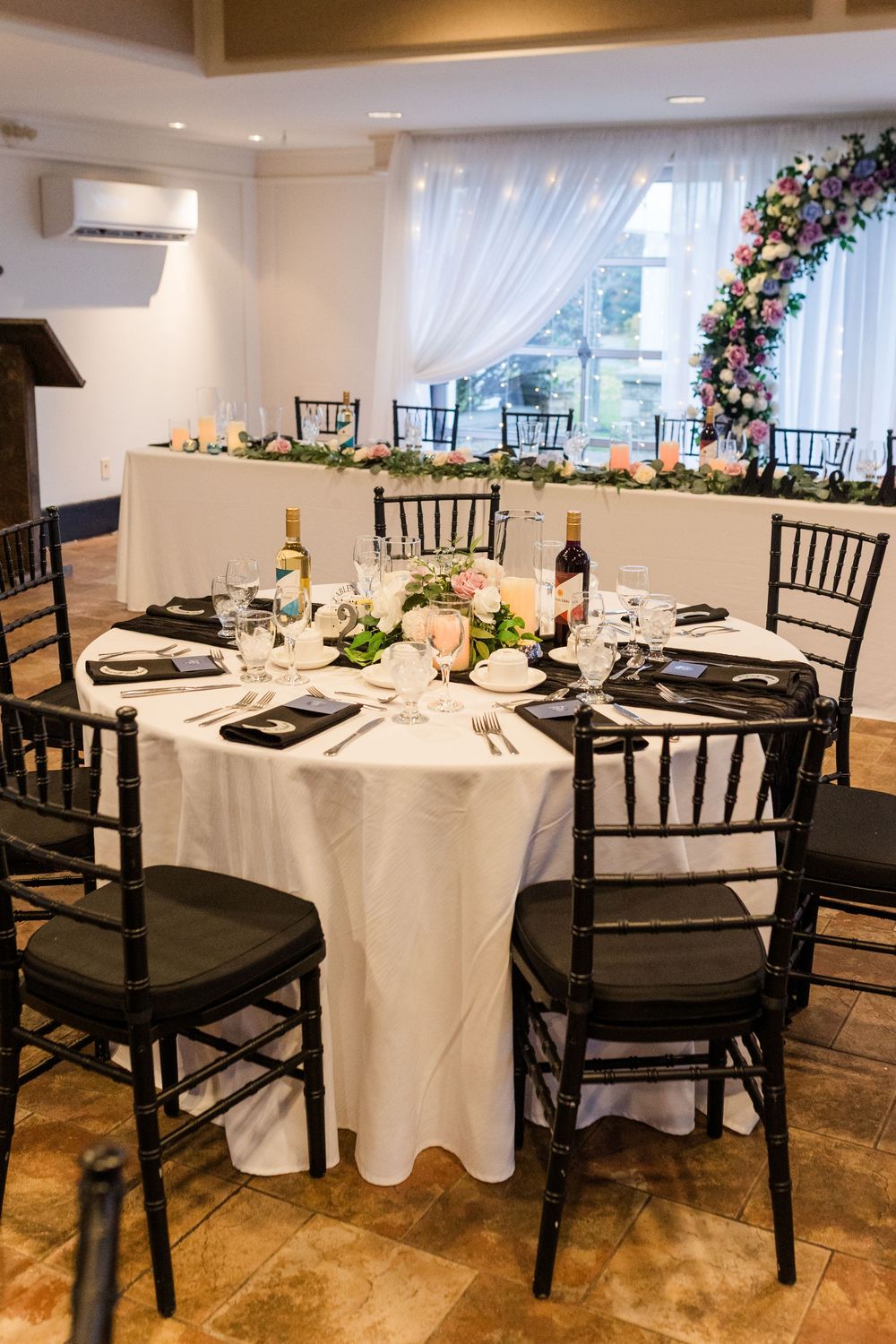 Elegant wedding reception setup with round tables featuring white linens, black chiavari chairs and floral arrangements.