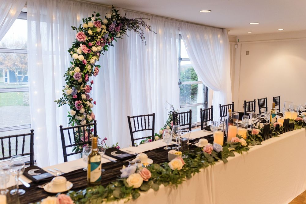 Elegant wedding reception venue with white curtains, dark Chiavari chairs, and floral arrangements featuring pink and white blooms.
