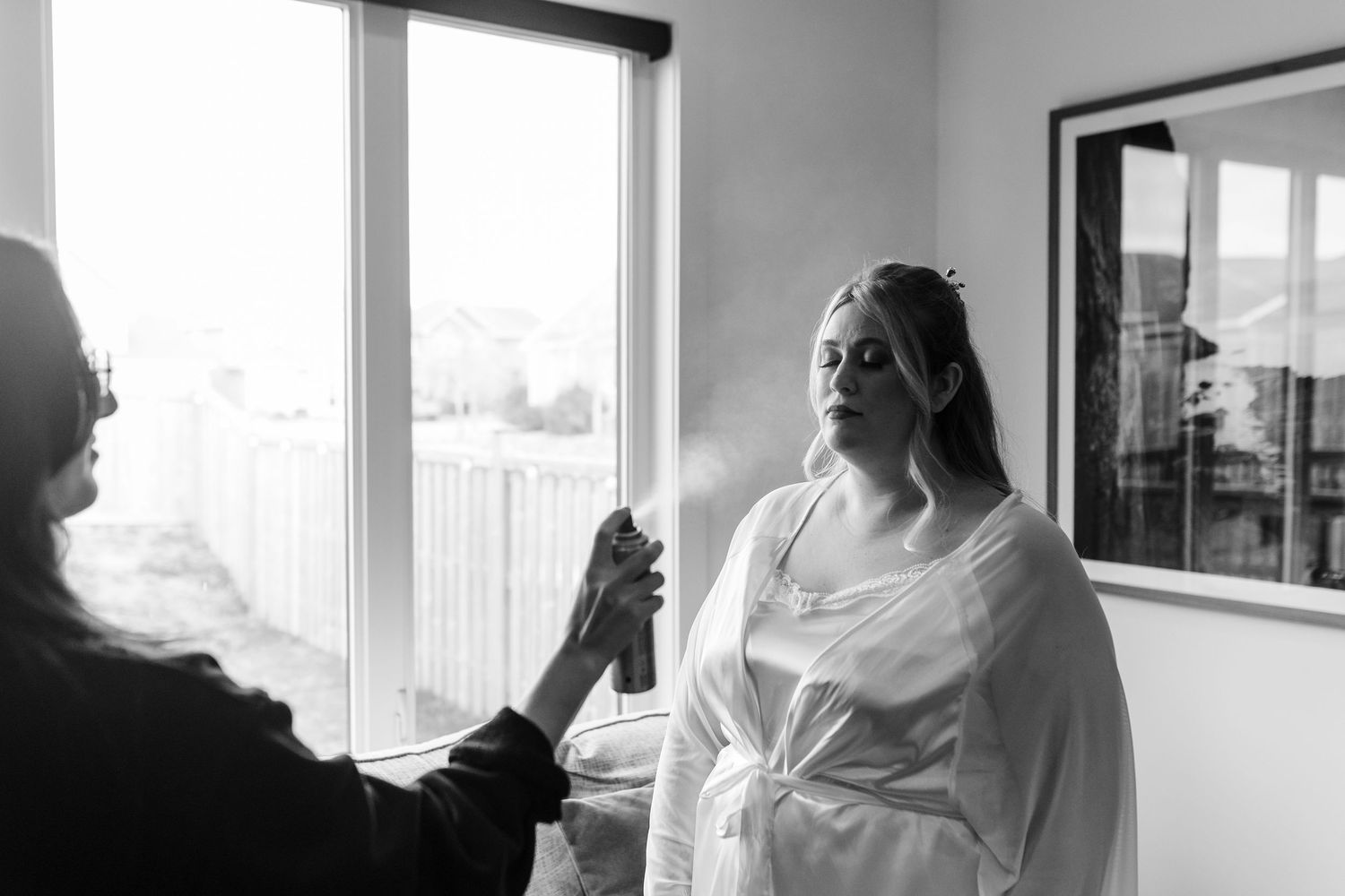 A person in a white robe gets ready in front of a window while someone helps with preparations.
