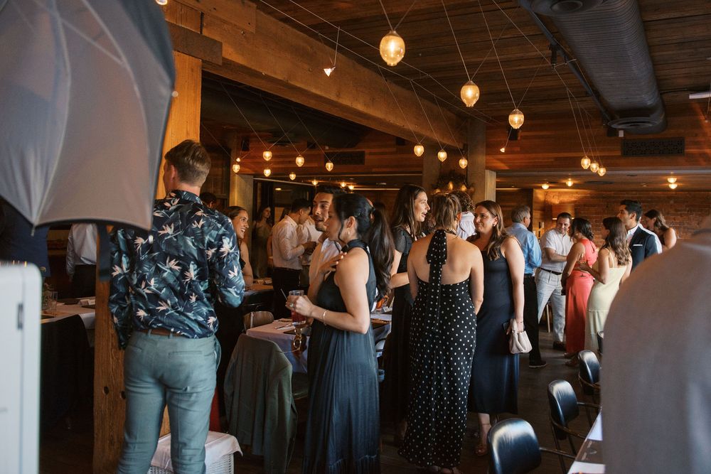 Wedding guests in formal attire gather inside a rustic venue with warm pendant lighting and exposed wooden beams.