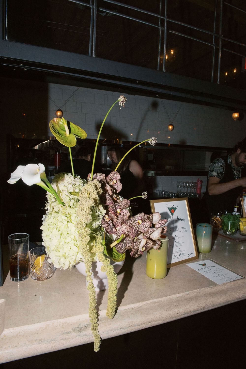 Elegant floral arrangements with hydrangeas and greenery line a dimly lit bar counter with cocktail glasses.