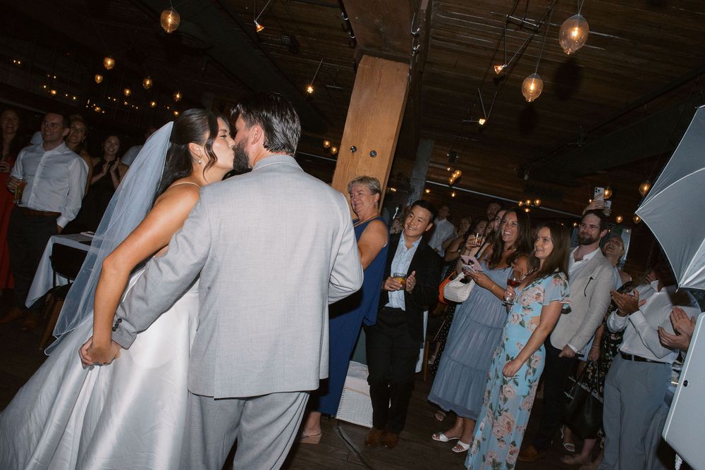 Wedding reception guests dancing and mingling in a rustic venue with wooden beams and string lights overhead.