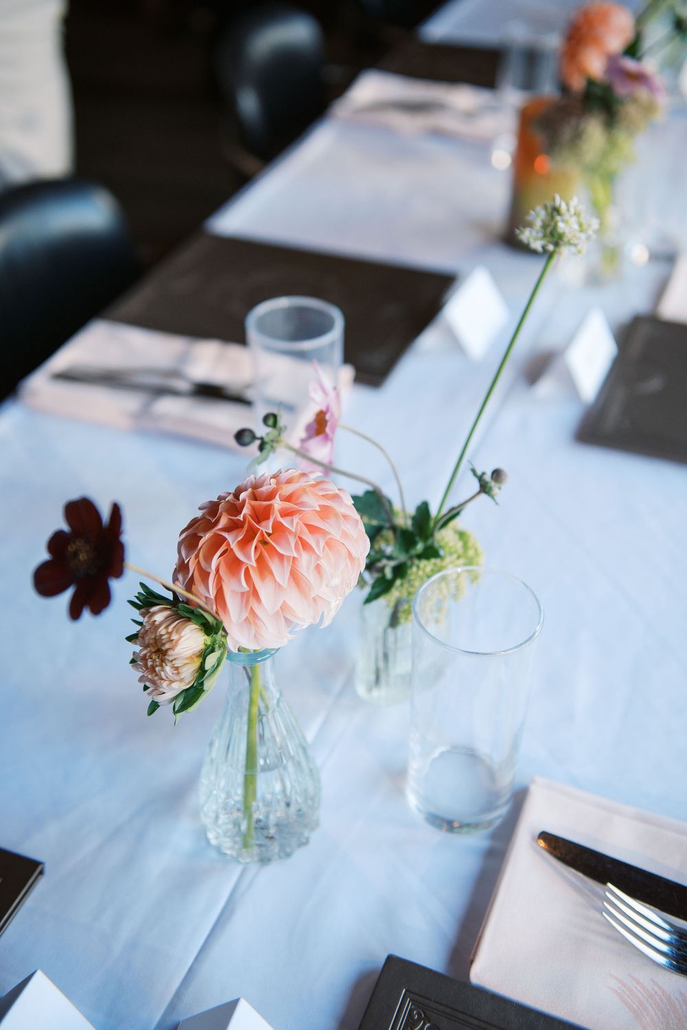 Delicate pink carnations and wildflowers arranged in small glass vases on a white tablecloth create an elegant table setting.