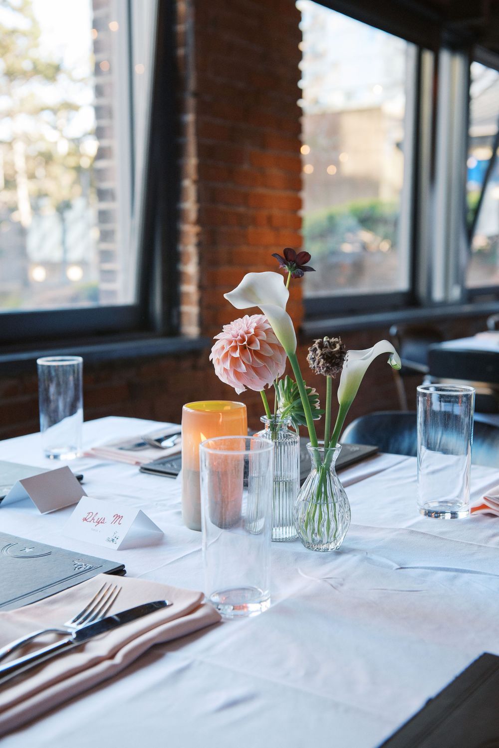Elegant restaurant table settings with white linens, pink calla lilies in glass vases, and natural lighting from windows.