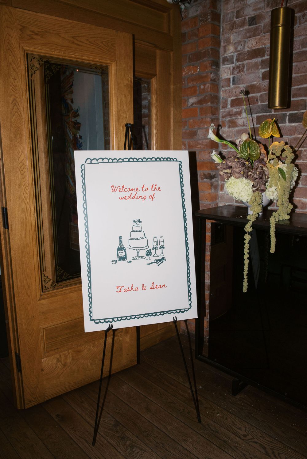 Welcome sign with floral decorations displayed on easel against brick wall at wedding venue.