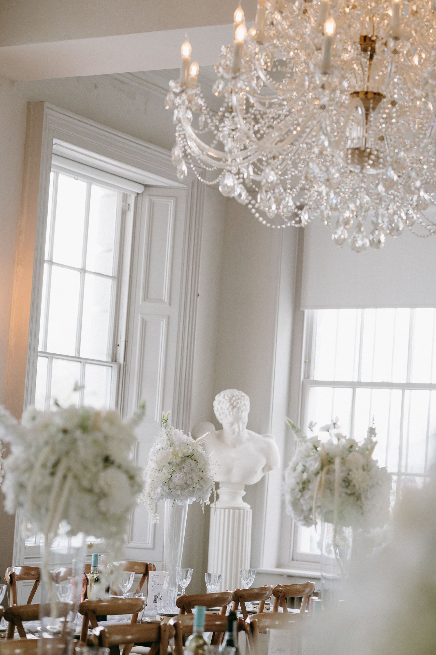 Elegant white wedding reception room with crystal chandeliers, tall windows, and white floral centerpieces on dining tables.