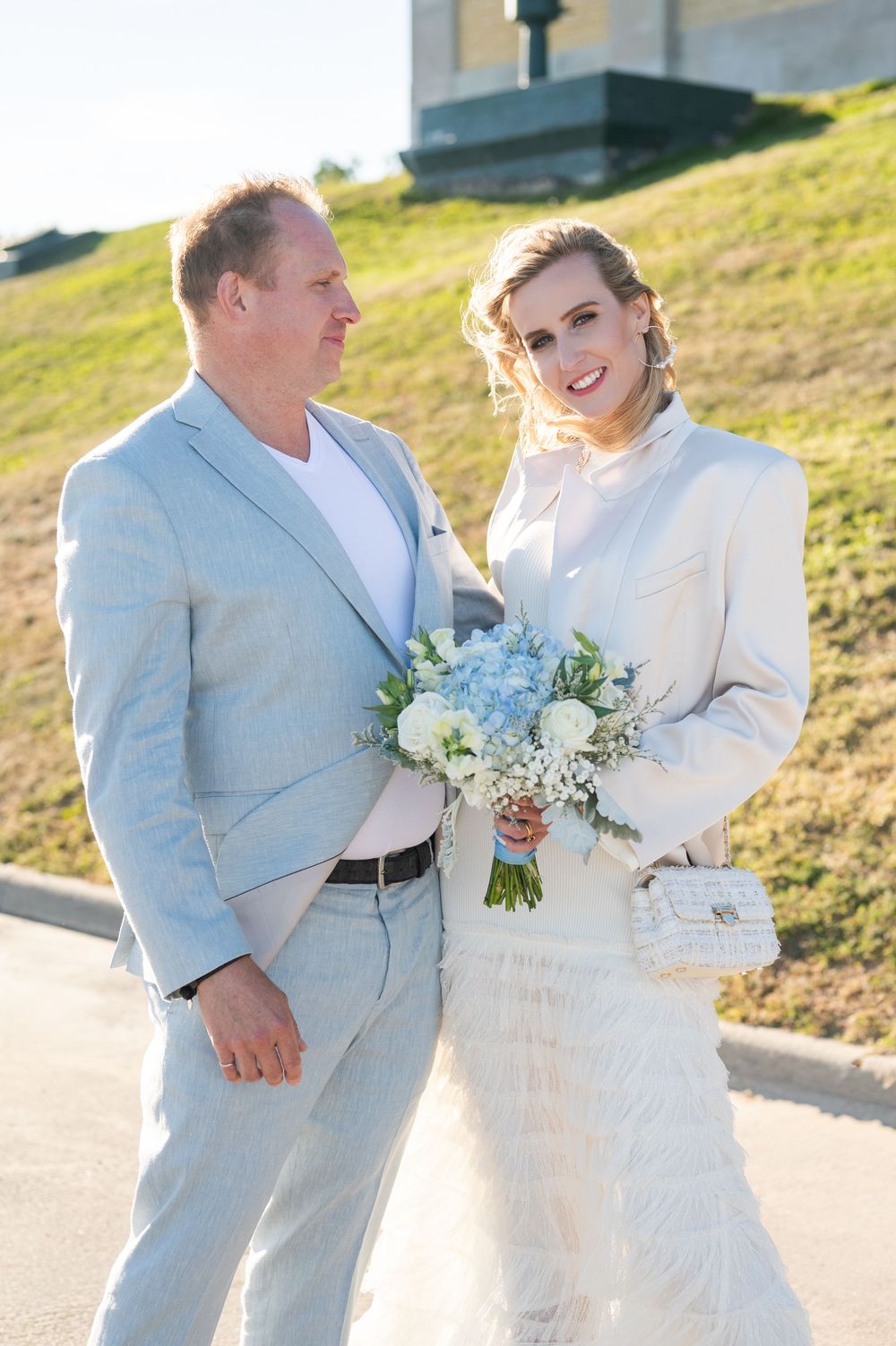 A joyful wedding couple embraces outdoors on a sunny day with blue hydrangea and white floral bouquet.