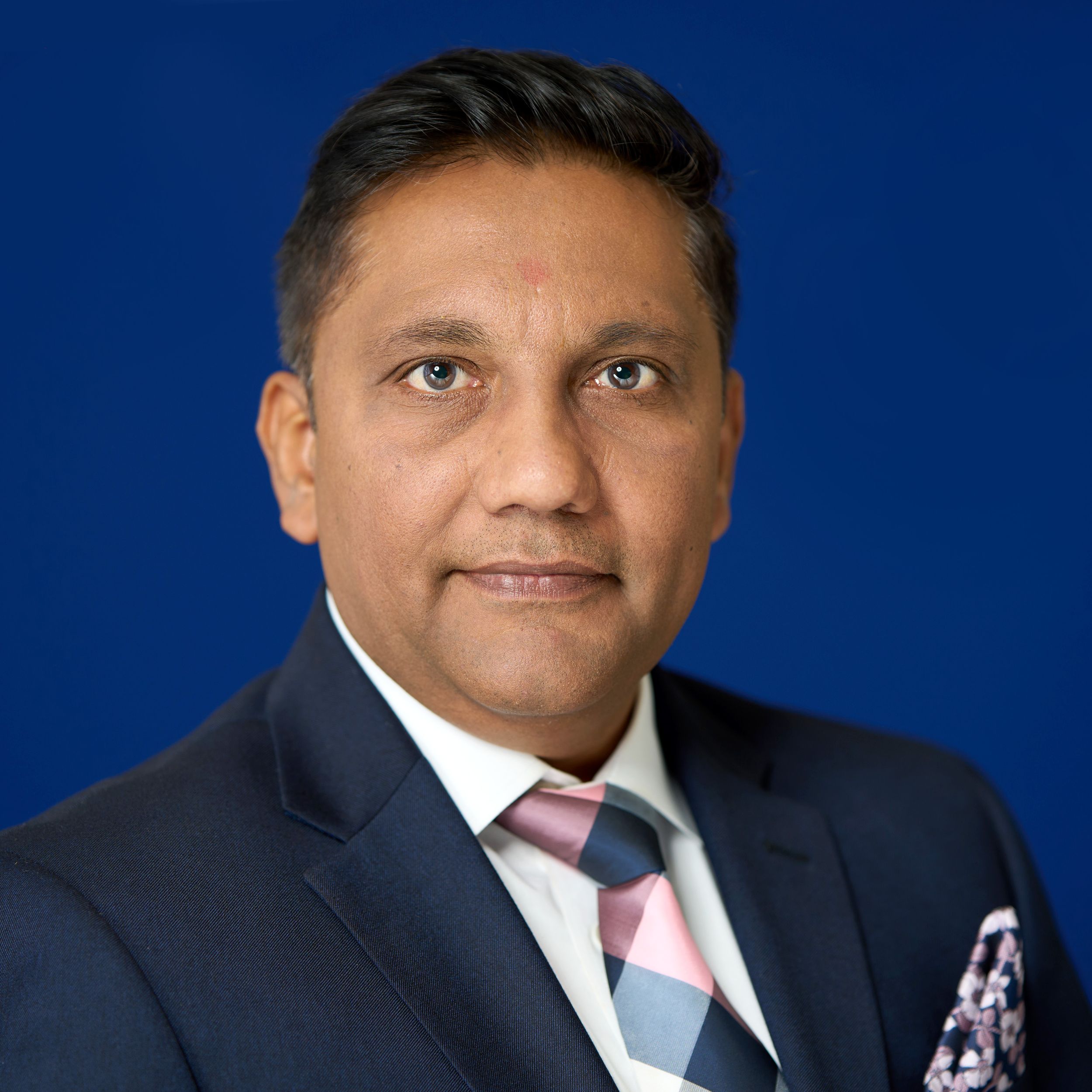 Professional headshot of someone in a dark suit and pink tie against a deep blue background.