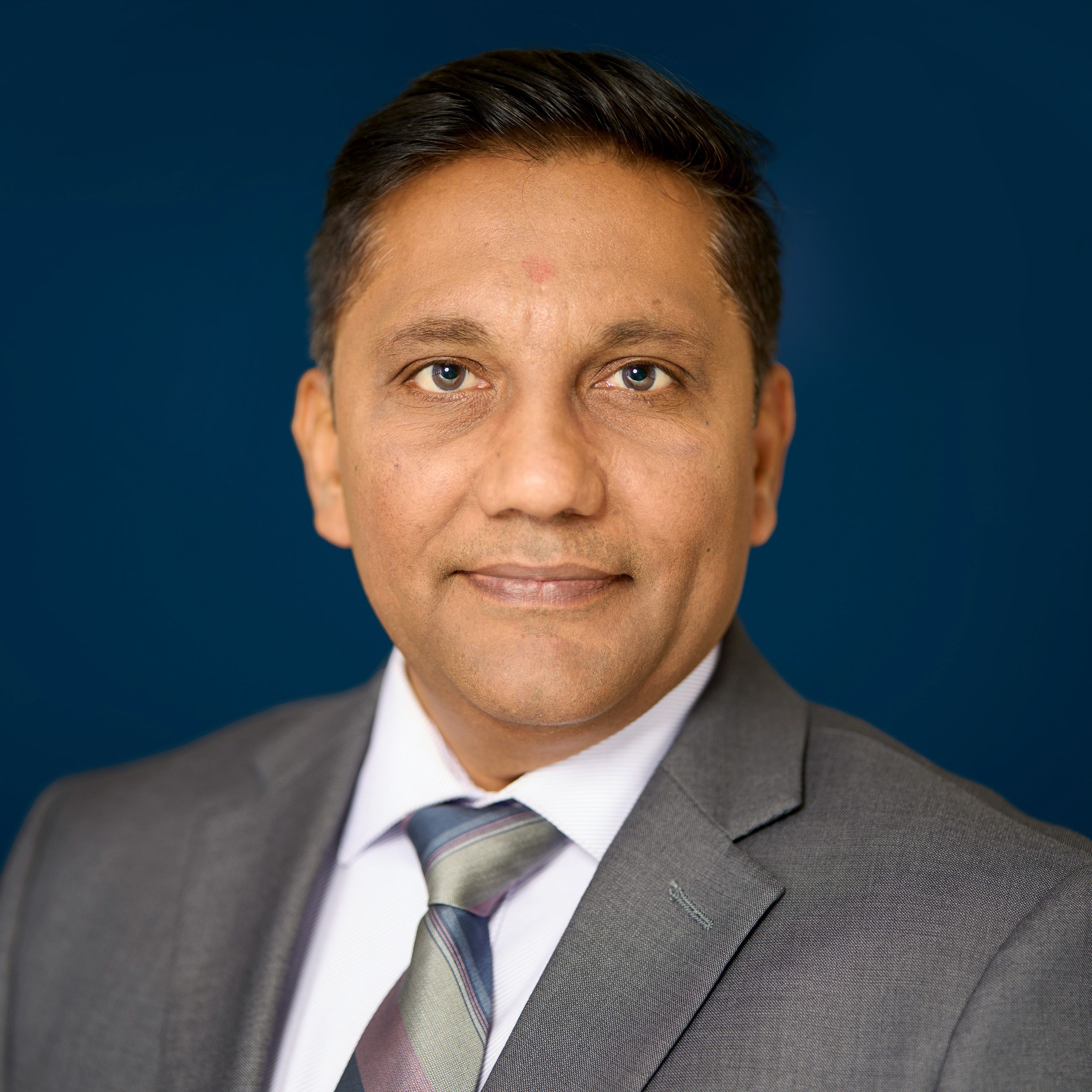 Professional headshot of person in grey suit and striped tie against dark blue background.