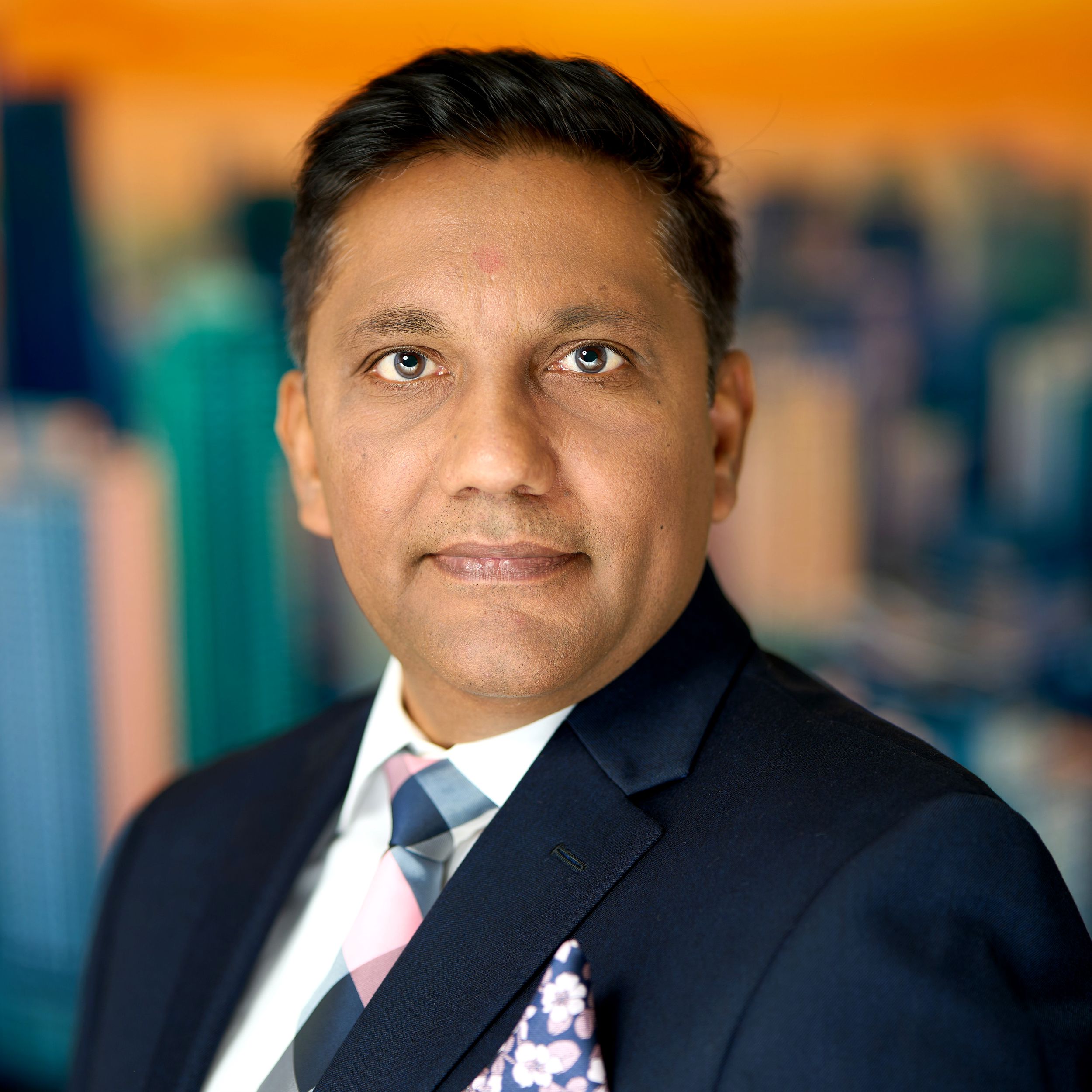 Professional business portrait of an executive in a navy suit and striped tie against a blurred orange background.