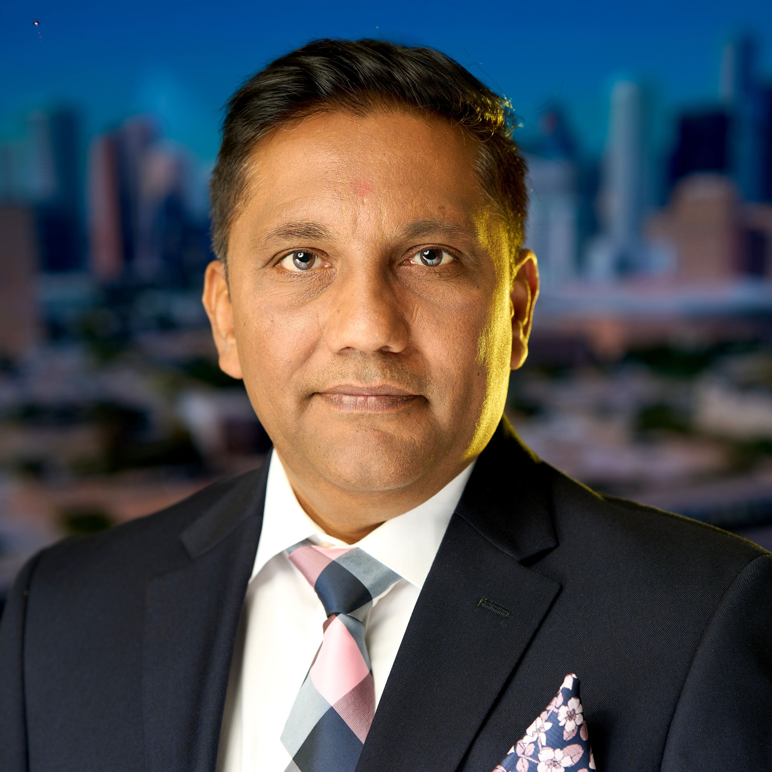 Professional headshot of someone in a dark suit and patterned pocket square against a blurred city background.