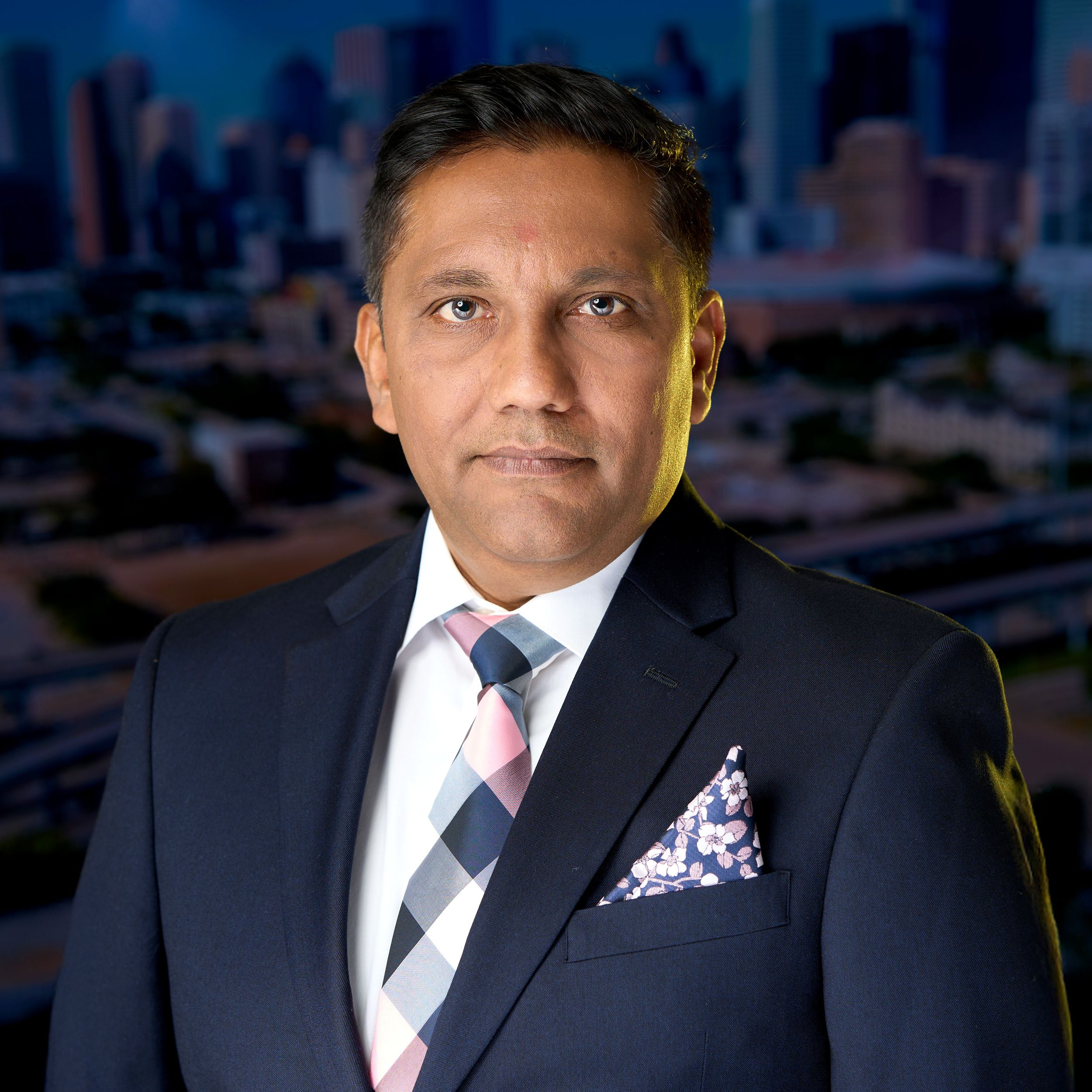 Professional wearing a dark suit with polka dot pocket square poses against blurred office background.