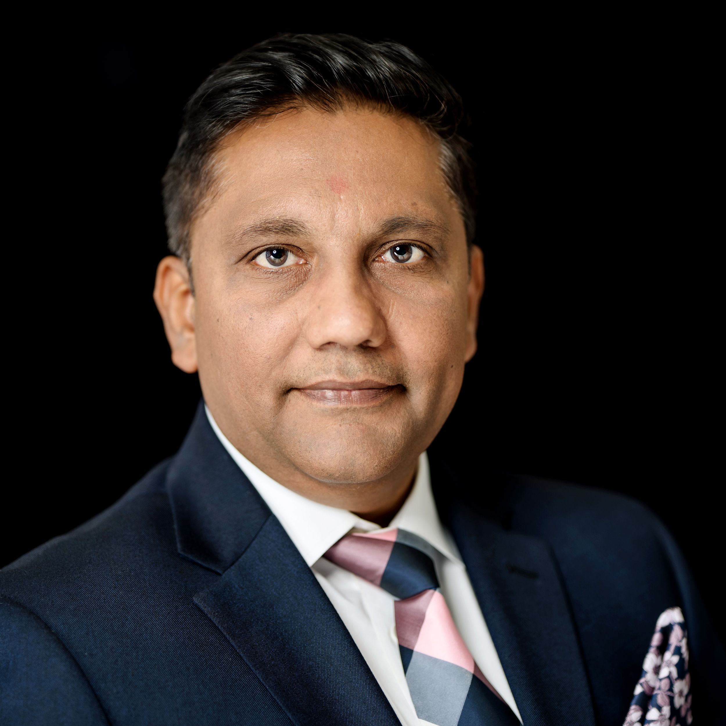 Professional headshot of someone in a dark suit with pink tie against black background.