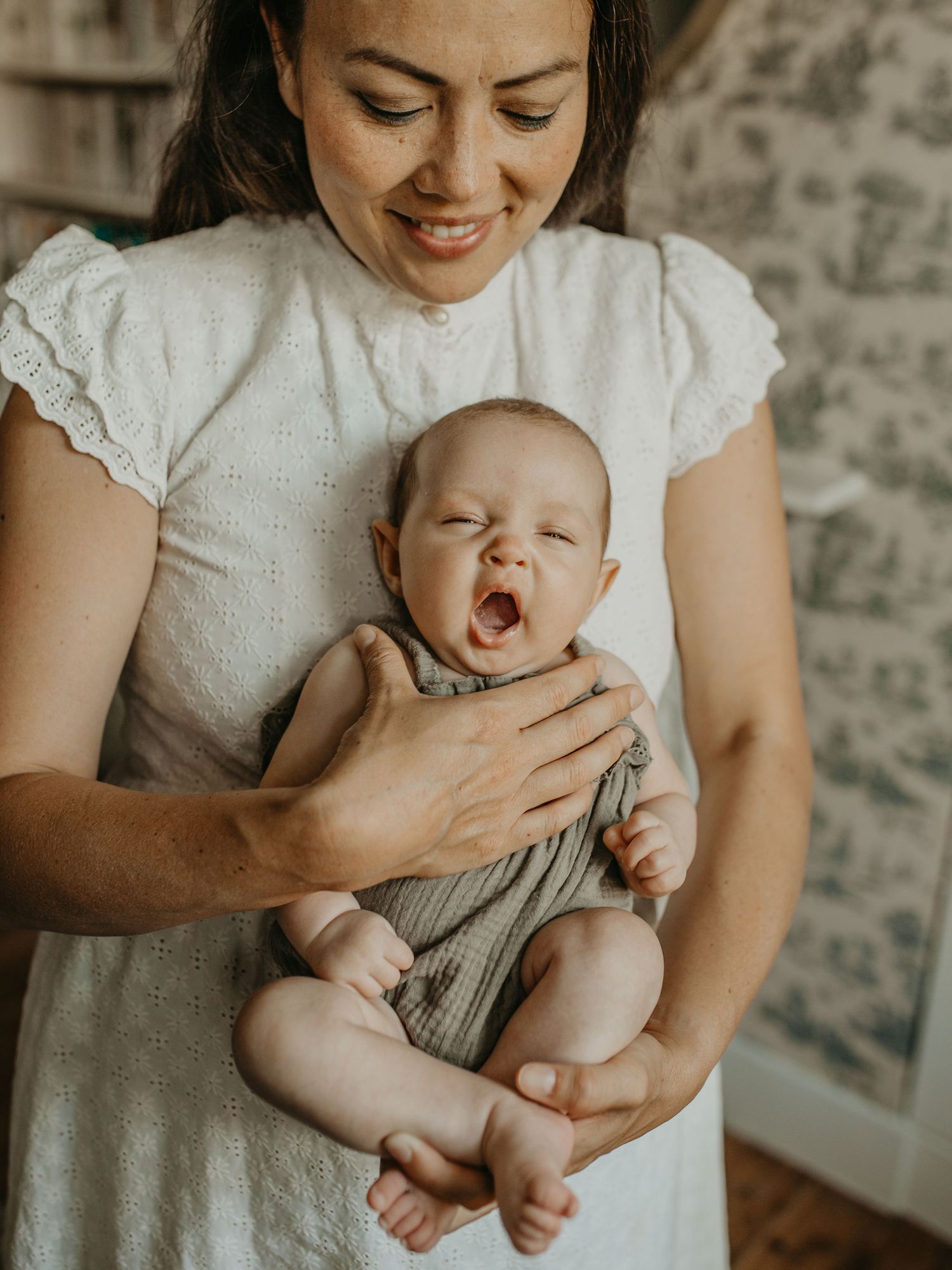 A tender moment shows a parent in a white dress holding a yawning baby wearing gray against a patterned wall background.