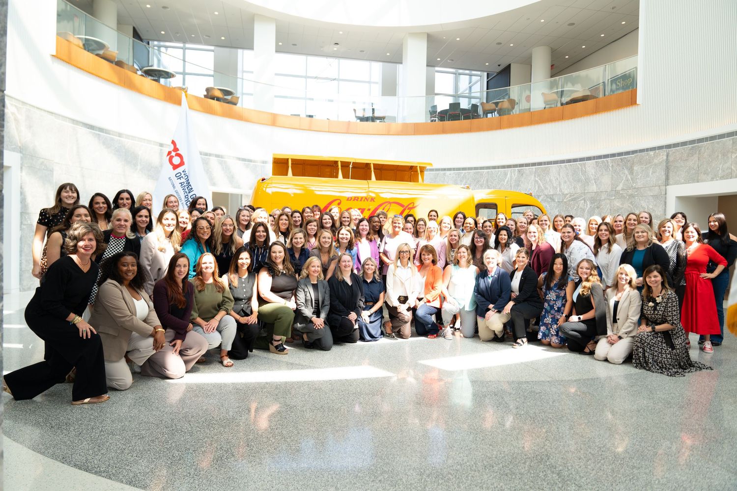 Large group of people seated in a curved formation inside a modern building with glass walls and natural lighting.