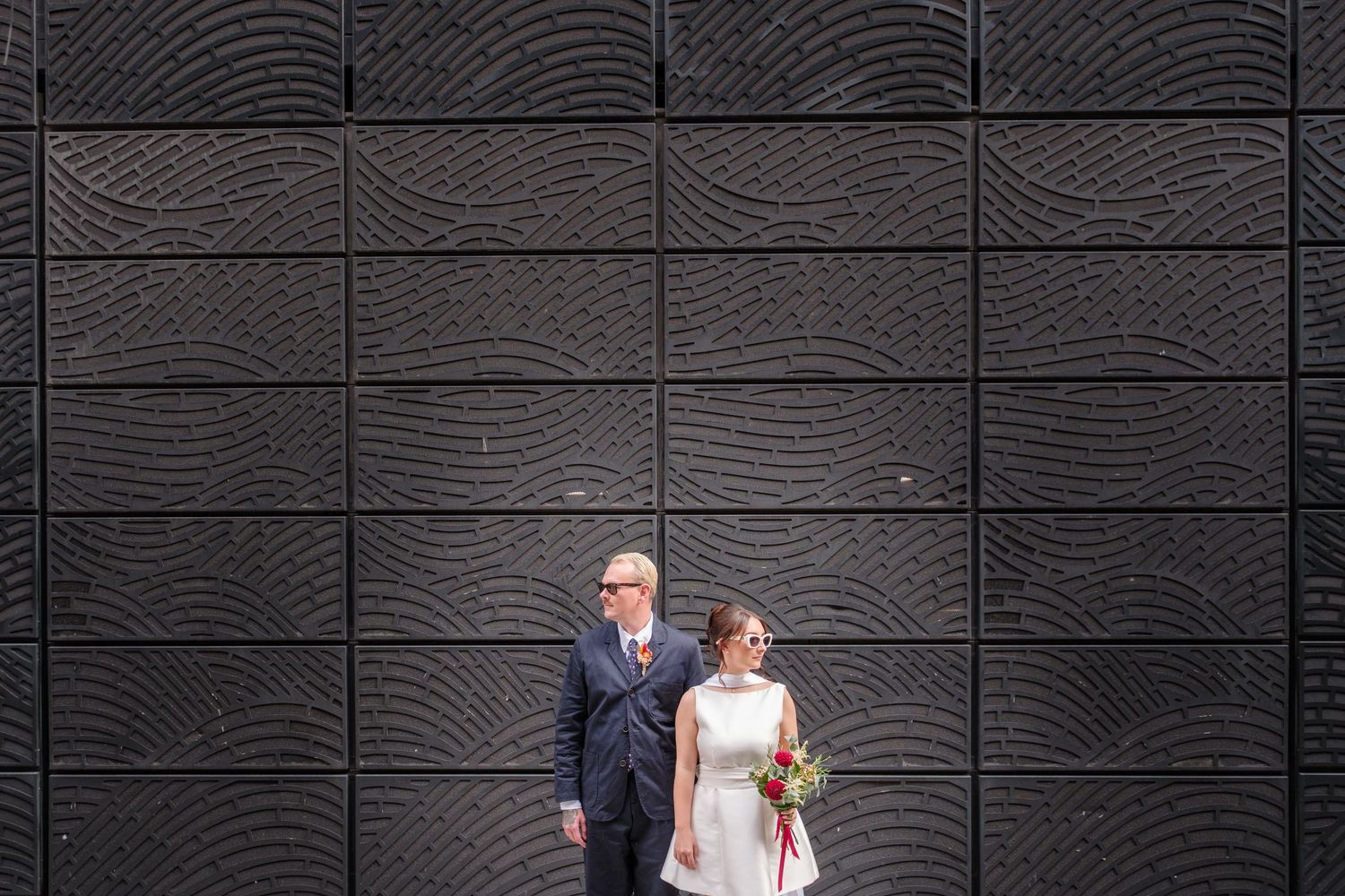 A couple in wedding attire stands against a dark textured wall with wood grain pattern.